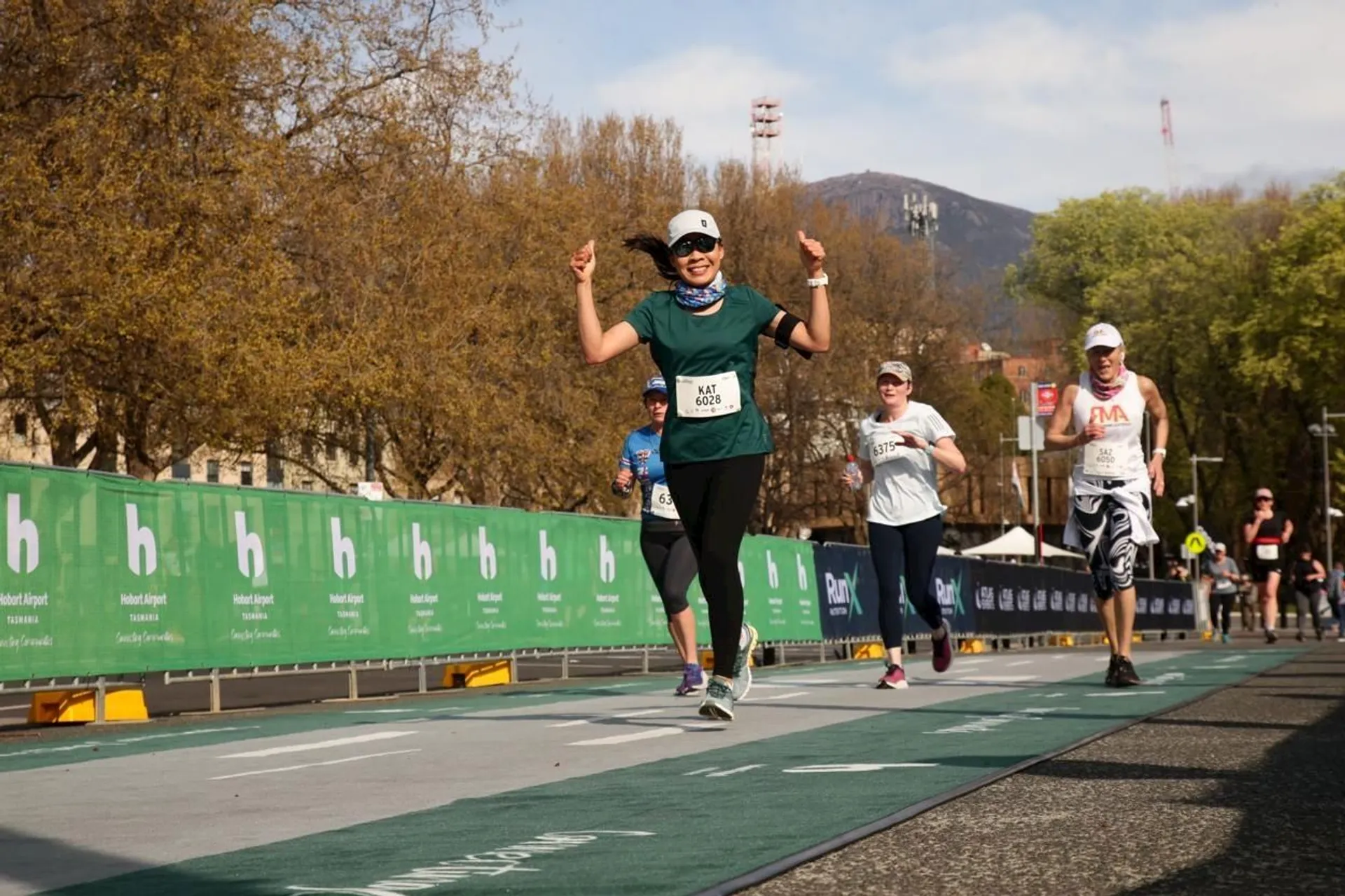 The image shows a group of people running in a race on a road. The runner in front is smiling and raising both arms in a celebratory gesture, wearing a green shirt and a cap. There are green barriers along the sides of the road with some spectators watching. The background features some trees and a mountain.