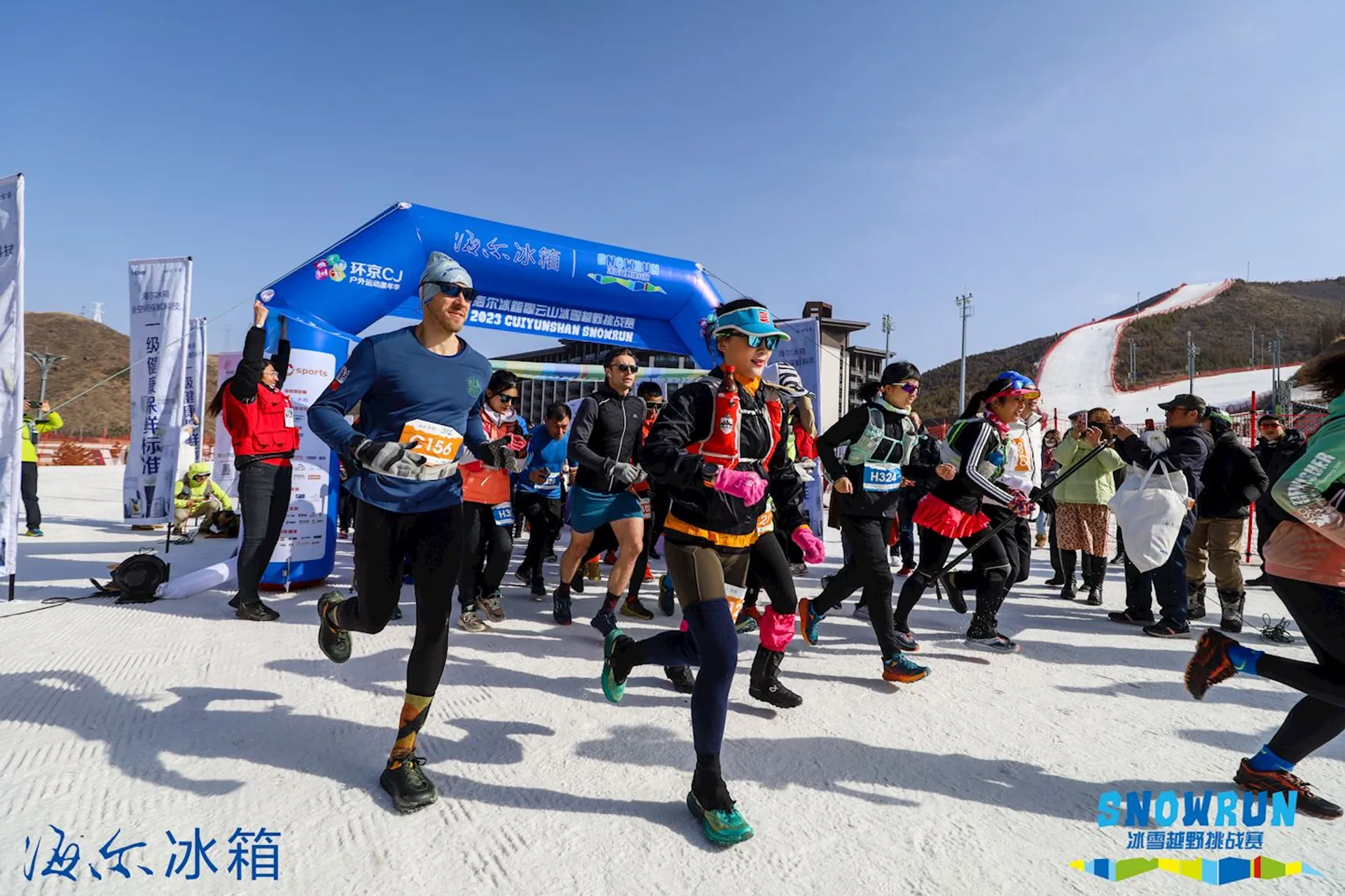The image shows a group of people participating in an outdoor running event. They are seen running past a start or finish line marked by an inflatable arch, which features some text and logos indicating sponsors or organizers. It appears to be a sunny day, and there's snow on the ground and hills in the background, suggesting that the event is taking place in a cold environment or during the winter season. The runners are dressed in active sportswear appropriate for the conditions, and they have numbers pinned to their clothing, which is typical for participants in a race. Everyone seems to be in motion and focused on their run.