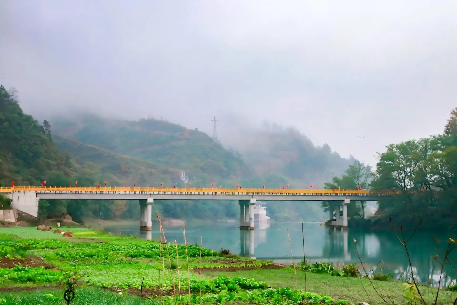 The image shows a scenic landscape with a bridge spanning a river. The bridge appears to have orange railings, and there are people walking on it. Lush green vegetation is in the foreground, and misty hills and trees are visible in the background. The overall atmosphere is serene and tranquil.