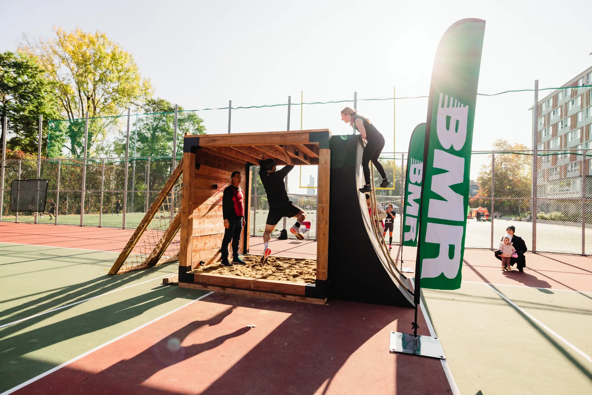 The image shows people interacting with a wooden obstacle course or skate ramp. One person is climbing over the top, while another appears to be jumping inside the wooden structure. There is a large banner with "BMR" written on it. The scene takes place on a sports court with a chain-link fence and trees in the background.