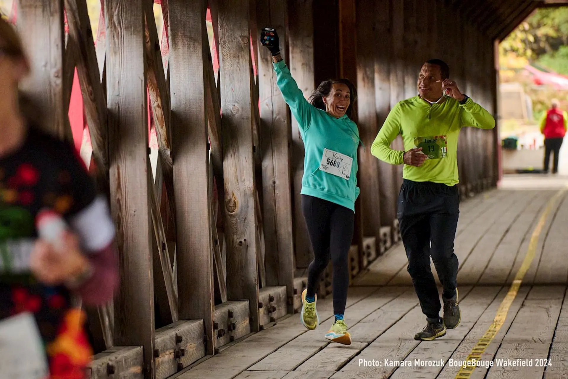 The image shows two people running side by side inside a covered wooden bridge. One person is enthusiastically holding up a smartphone, and both are wearing numbered race bibs. They appear to be participating in an organized running event.