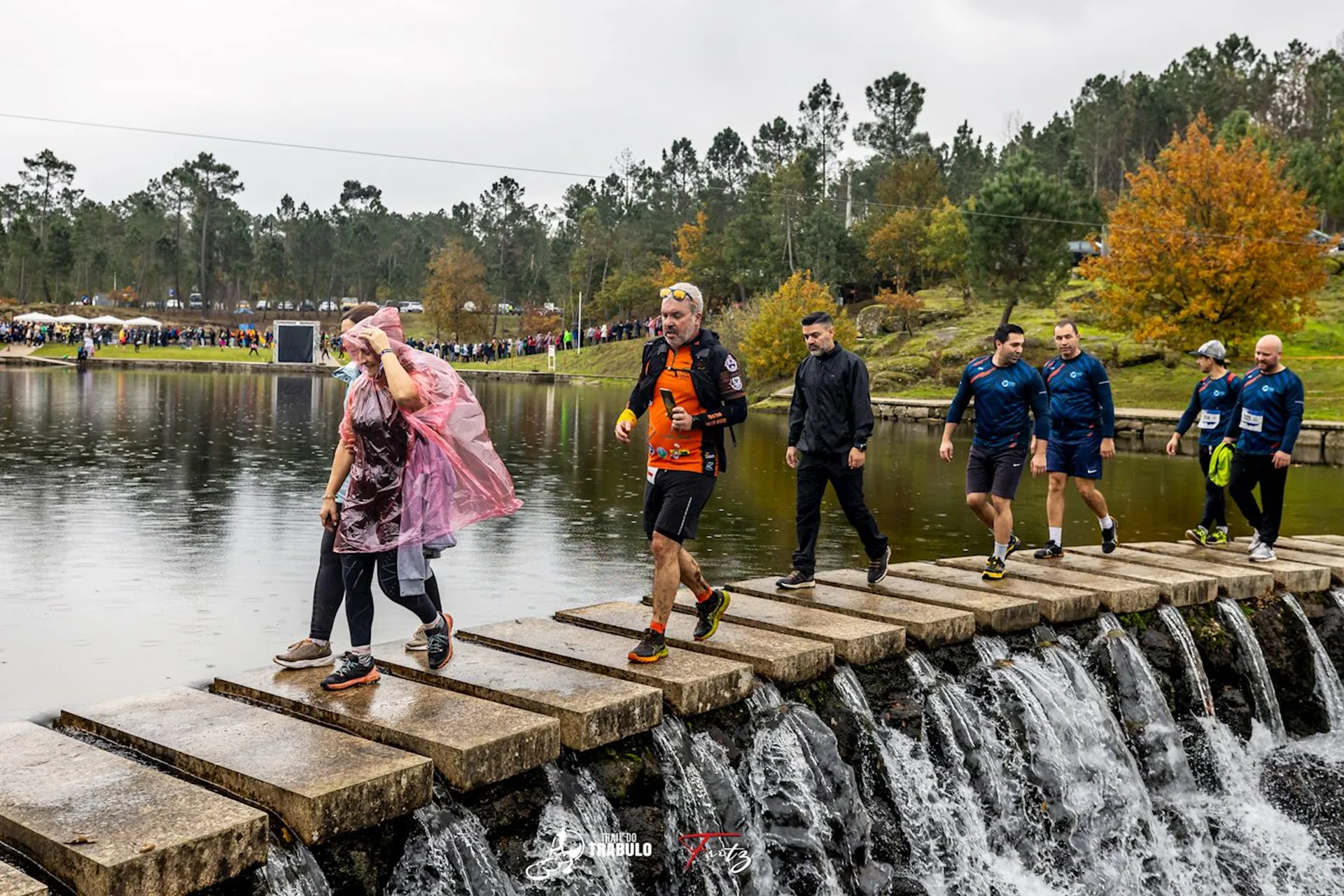 The image shows a group of people participating in an outdoor event, crossing a stepping stone bridge over flowing water. Some participants are wearing running attire and appear to be mid-event. The setting is natural, with trees and a body of water visible in the background. The atmosphere seems lively, indicating a race or fun run event near a park with spectators in the background.