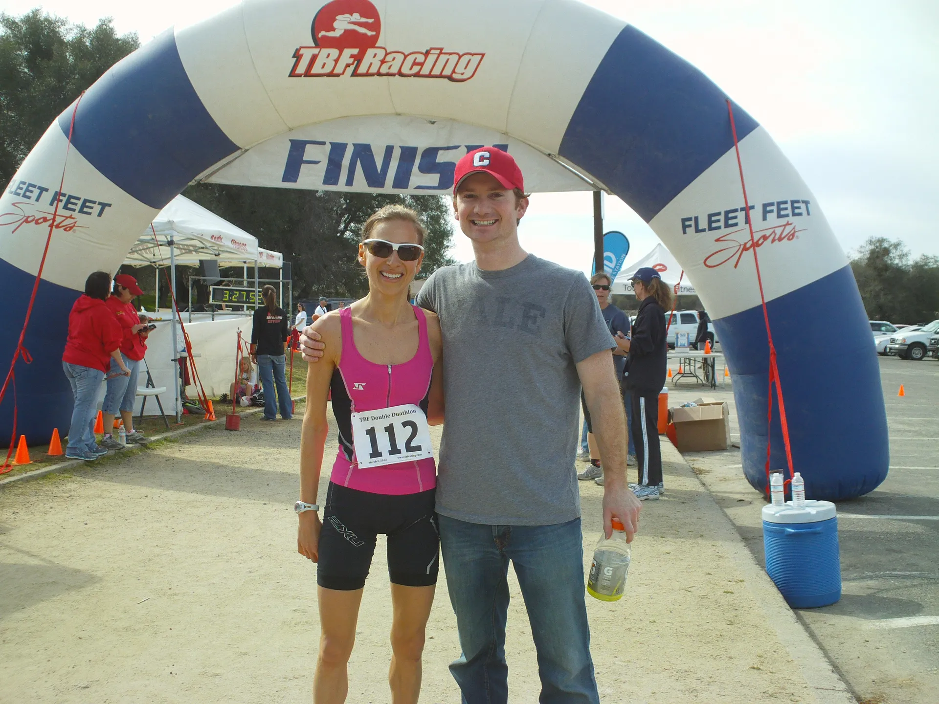 The image shows two people standing under an inflatable arch with the words "TBF Racing" and "FINISH" on it, indicating that they are at the finish line of a race or sporting event. The person on the left appears to be a female athlete wearing a numbered racing bib (112) and fitness attire, while the person on the right is a male wearing a casual t-shirt and a baseball cap with a letter "C" on it. They both appear to be smiling and posing for the photo with a background of trees and a clear sky. The environment suggests an outdoor event organized by TBF Racing, and several event-related structures like cones and a tent with the "Fleet Feet Sports" branding are visible, which may indicate a