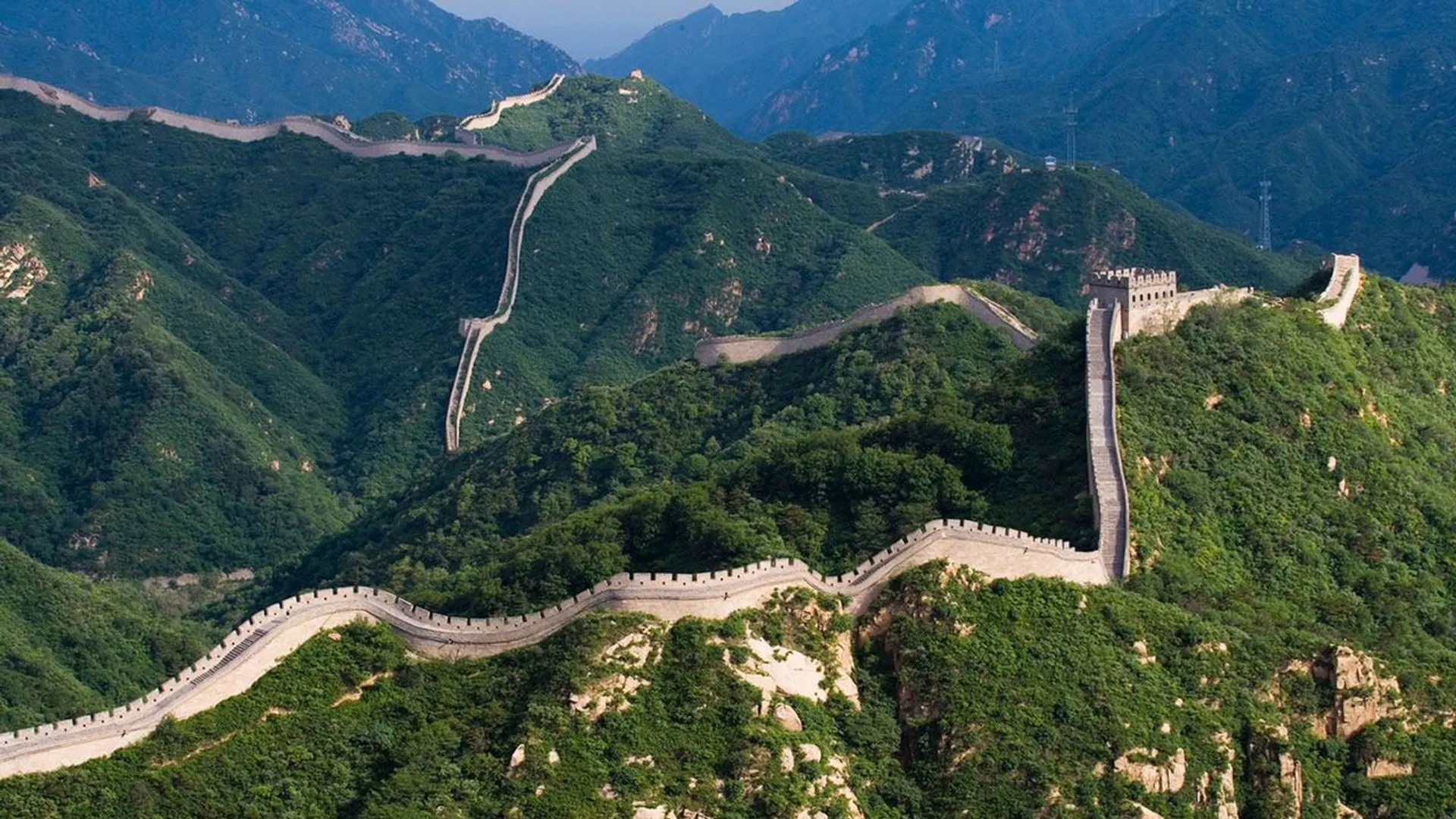 This image shows the Great Wall of China winding through hilly terrain. The wall is a series of fortifications made of stone and other materials, stretching across mountains and lush green landscapes.