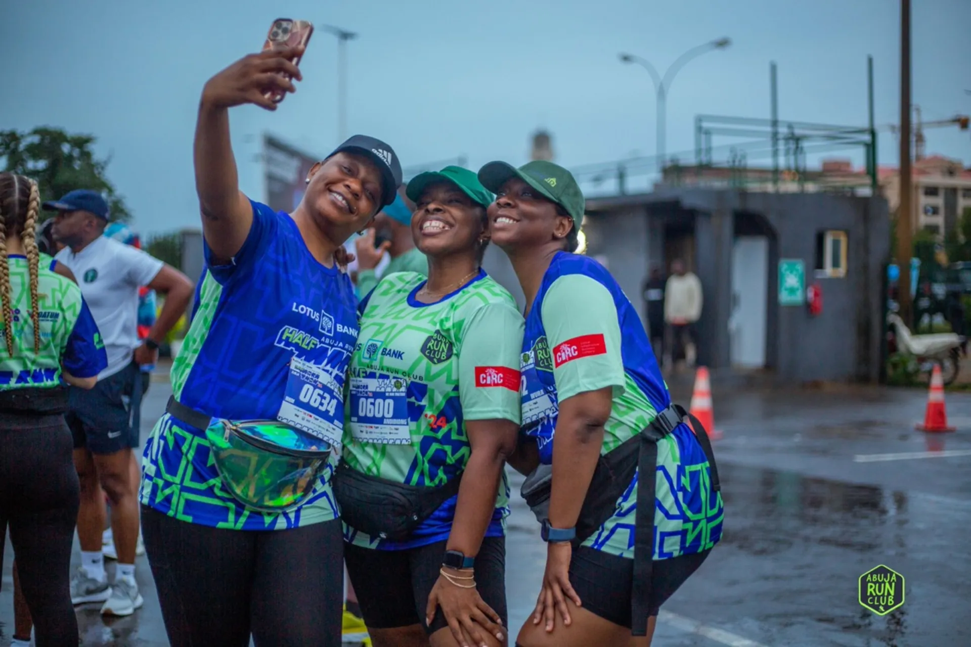 The image shows three people taking a selfie together. They are wearing athletic gear and race bibs, likely indicating participation in a running event. The setting appears to be outdoors, possibly near a race starting or finishing area. There are other individuals and some urban elements like buildings and signs visible in the background.