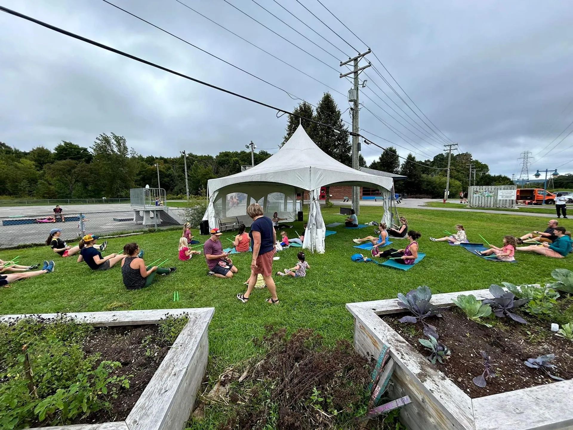 The image shows a group of people participating in an outdoor exercise or yoga class on a grass field. There is a white tent in the background, and some attendees are using yoga mats. In the foreground, there are raised garden beds with plants. The setting has a casual and community-oriented atmosphere.
