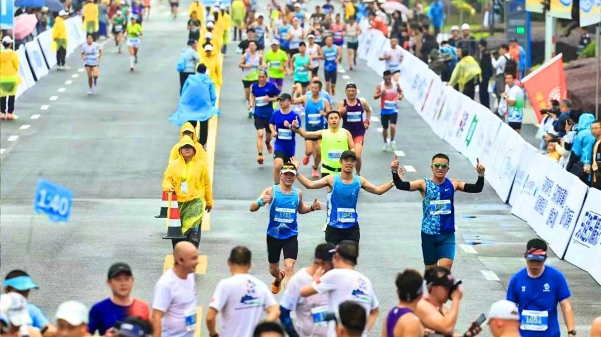 The image depicts a marathon event with numerous runners participating. Participants are running down a street with race bibs on their shirts. Some are raising their arms enthusiastically. The street is lined with barricades, and there are spectators and event staff along the sides. Some people are wearing rain gear, suggesting it might be raining or that rain is expected.