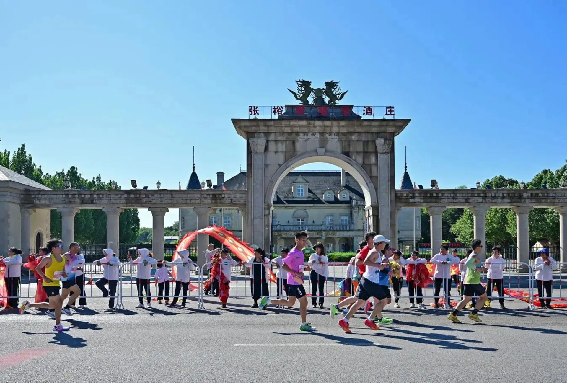 This image shows a group of people running a marathon or race. They are passing by an archway with a building and flags in the background. Spectators are standing on the side, cheering them on. The scene is in a large outdoor area with clear skies.