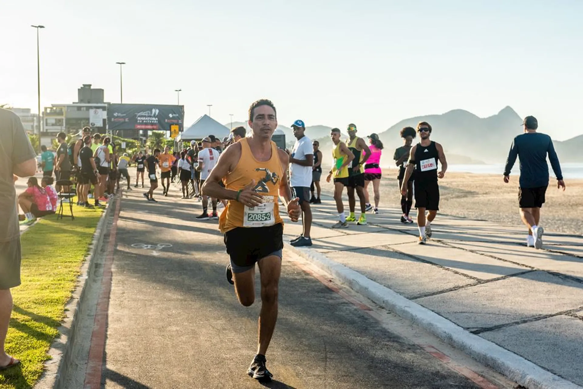 The image depicts a running event, likely a marathon or a race, with a focus on a male runner in the foreground, who is numbered 2085 and appears to be running at a faster pace than the others. He seems to be putting in a great effort, as indicated by his intense expression and running posture.

In the background, there are many other participants walking or jogging at a more relaxed pace. The setting appears to be a coastal road since there is a beach visible on the left and mountains in the far distance, which suggests the event might be taking place in a scenic location often chosen for such races. The weather looks sunny and clear, and it seems to be a well-organized event, with barriers set up along the side of