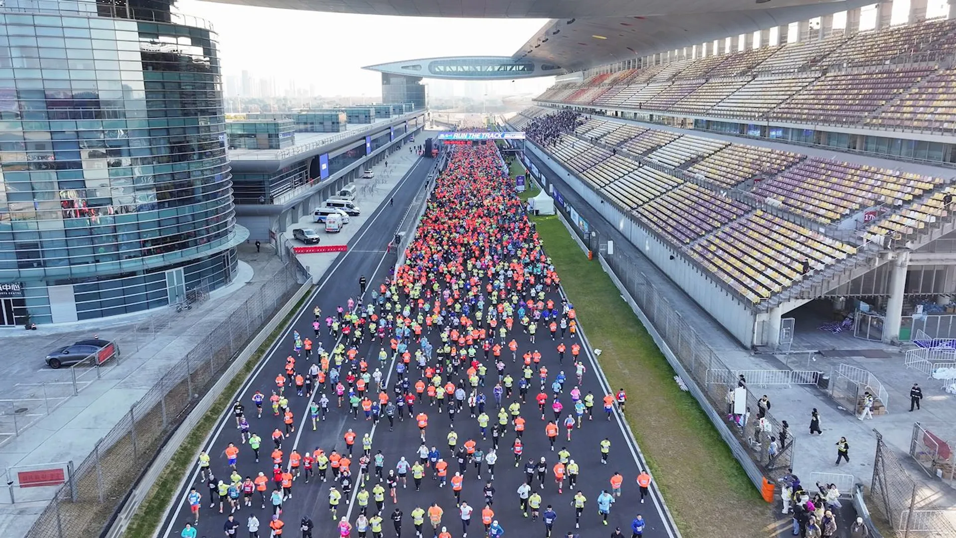 This image shows a large group of people participating in a marathon or running event on a track. The setting appears to be a stadium or racing circuit, with grandstands on one side and a large building on the other. The participants are wearing brightly colored clothing as they run along the track.