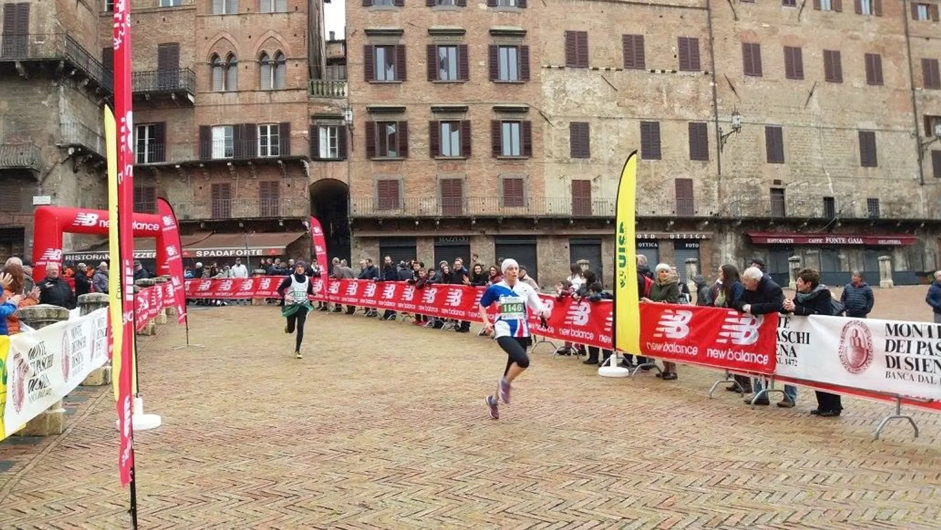 The image shows a runner crossing the finish line at a running event. You can see barricades with sponsor names such as New Balance lining the path, and there are spectators on either side, some seated and some standing, watching the runner. The location appears to be a historic square with old brick buildings surrounding it, which suggests that the race could be held in a city with a rich architectural heritage. The ground is paved with patterned tiles or bricks, adding to the aesthetic of the place. It looks like an overcast day, and the setting gives a sense of a community or city event that attracts both participants and spectators.