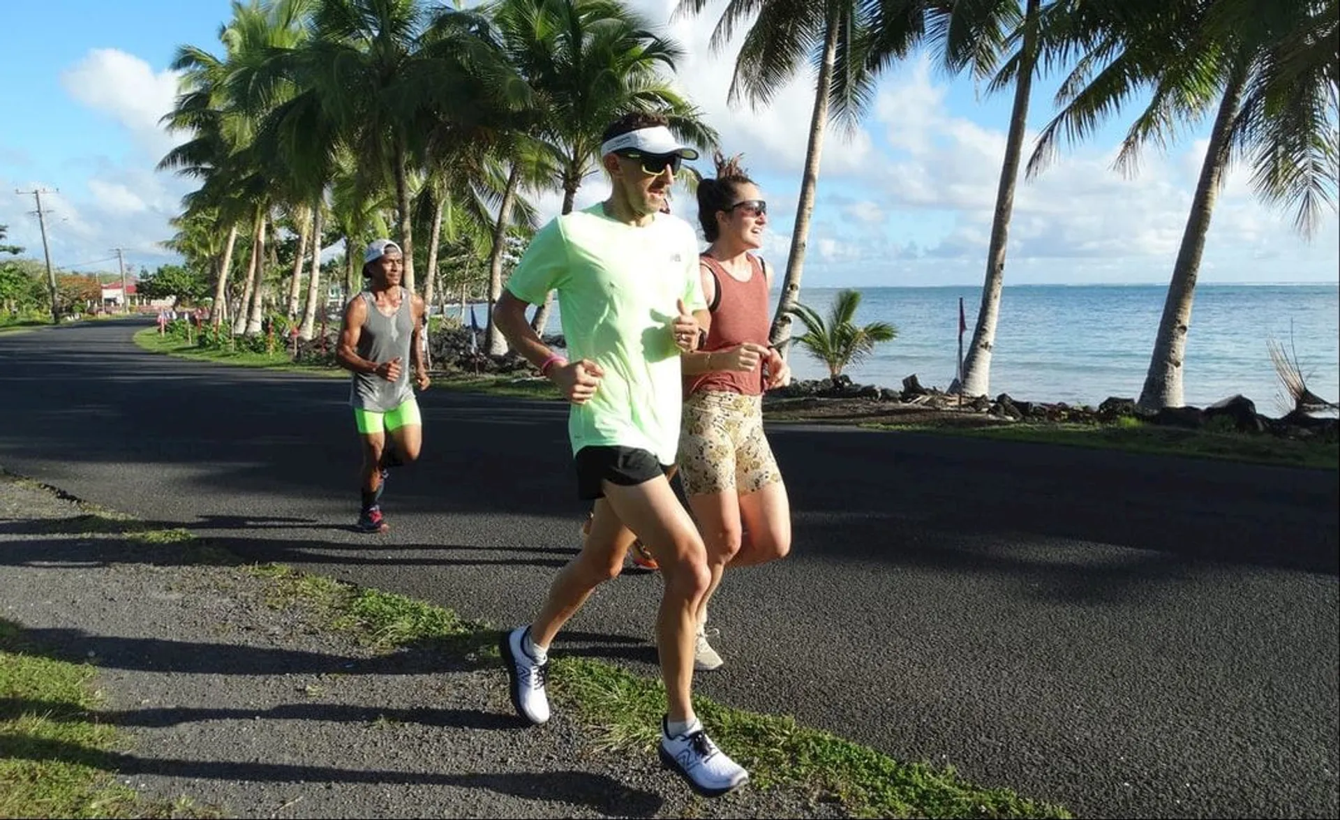 The image shows people jogging along a coastal road. They are surrounded by palm trees and it appears to be a sunny day. The ocean is visible on one side, and the atmosphere looks relaxed and tropical.