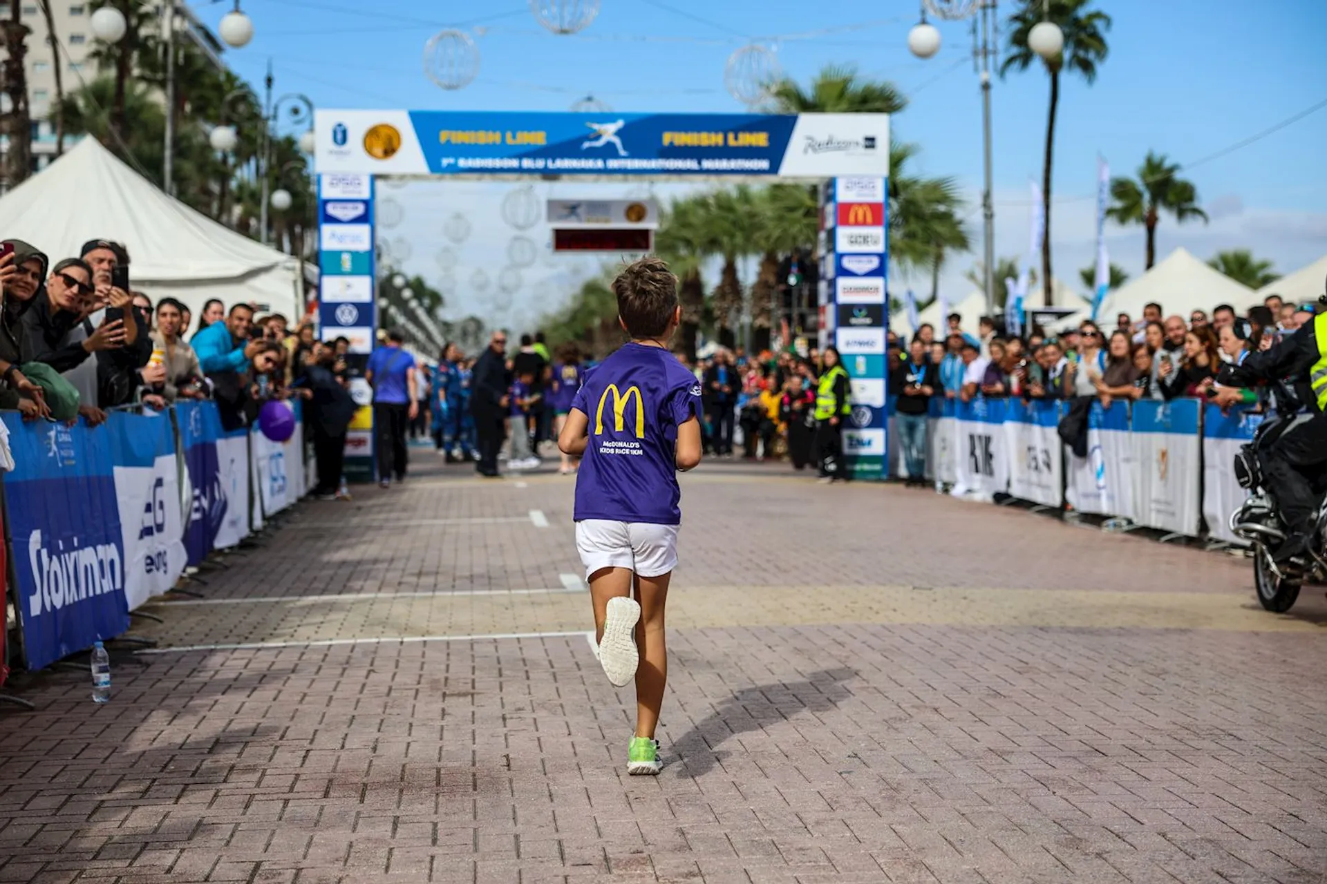 The image shows a young runner approaching the finish line of a marathon or race event. The runner is wearing a blue shirt with a large yellow letter on the back and white shorts. There are people on both sides cheering and taking photos as the runner approaches the finish line. Various banners and signs are visible around the finish area.