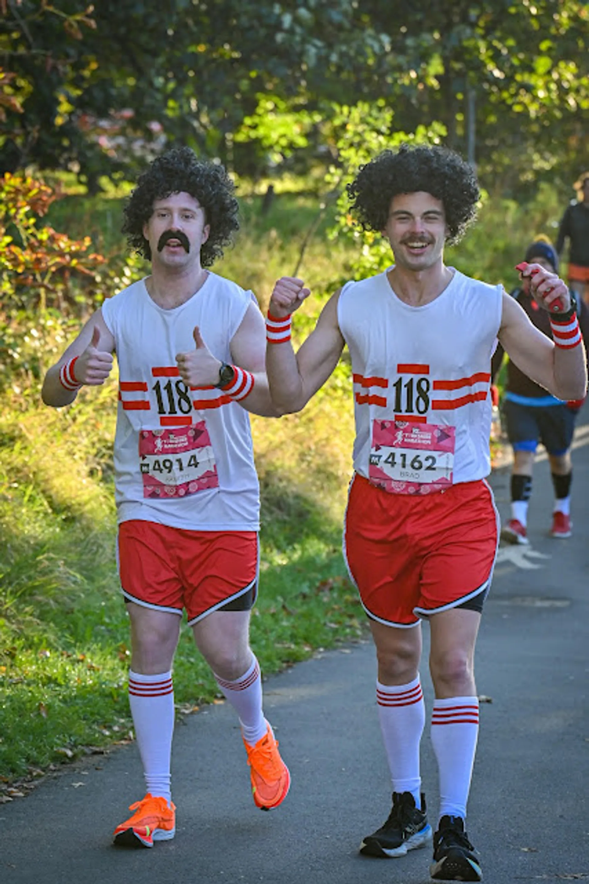 In the image, there are two individuals dressed in matching costumes running in what appears to be a road race or marathon. They are wearing white shirts with the number "118" printed in red, along with red shorts, white socks with two red stripes at the top, and athletic shoes. Both are sporting large, curly black wigs and fake black mustaches, which suggests they might be dressed up as a well-known character or theme for a fun run or charity event. They are giving thumbs-up gestures, indicating they are in good spirits and enjoying the event.