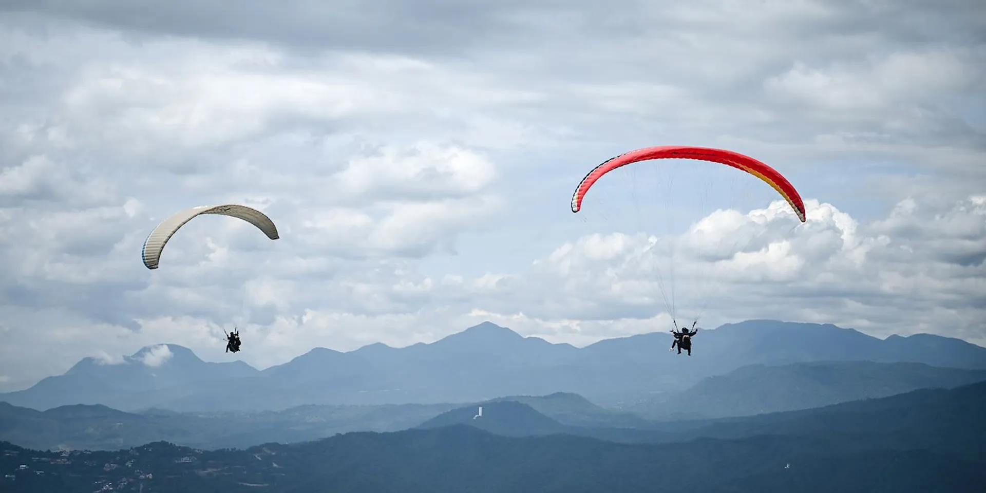 The image depicts two paragliders soaring in the sky. The paraglider on the left has a predominantly white wing with some patterns or colors toward the center, while the one on the right has a vibrant red wing, making it stand out against the sky. Both paragliders are equipped with harnesses and are suspended beneath their respective parachutes, which allow them to glide through the air. In the background, you can see a range of mountains or hills providing a scenic backdrop, with some clouds scattered across the sky. It's a serene image that conveys a sense of freedom and the thrill of flying.