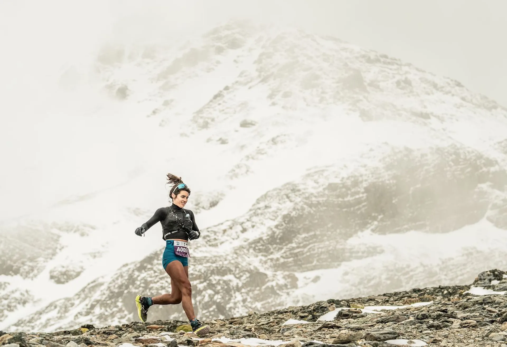 The image shows a person running on a mountainous and rocky terrain. The background features snow-covered mountains. The runner is dressed in athletic gear suitable for cold weather. The setting appears to be challenging and rugged.