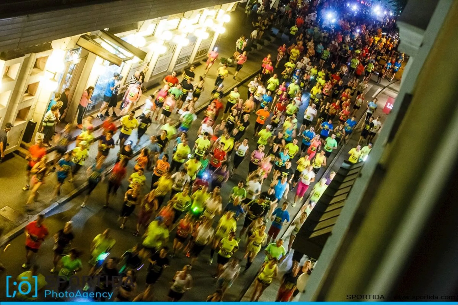 The image depicts a nighttime event where a large group of people wearing colorful clothing is