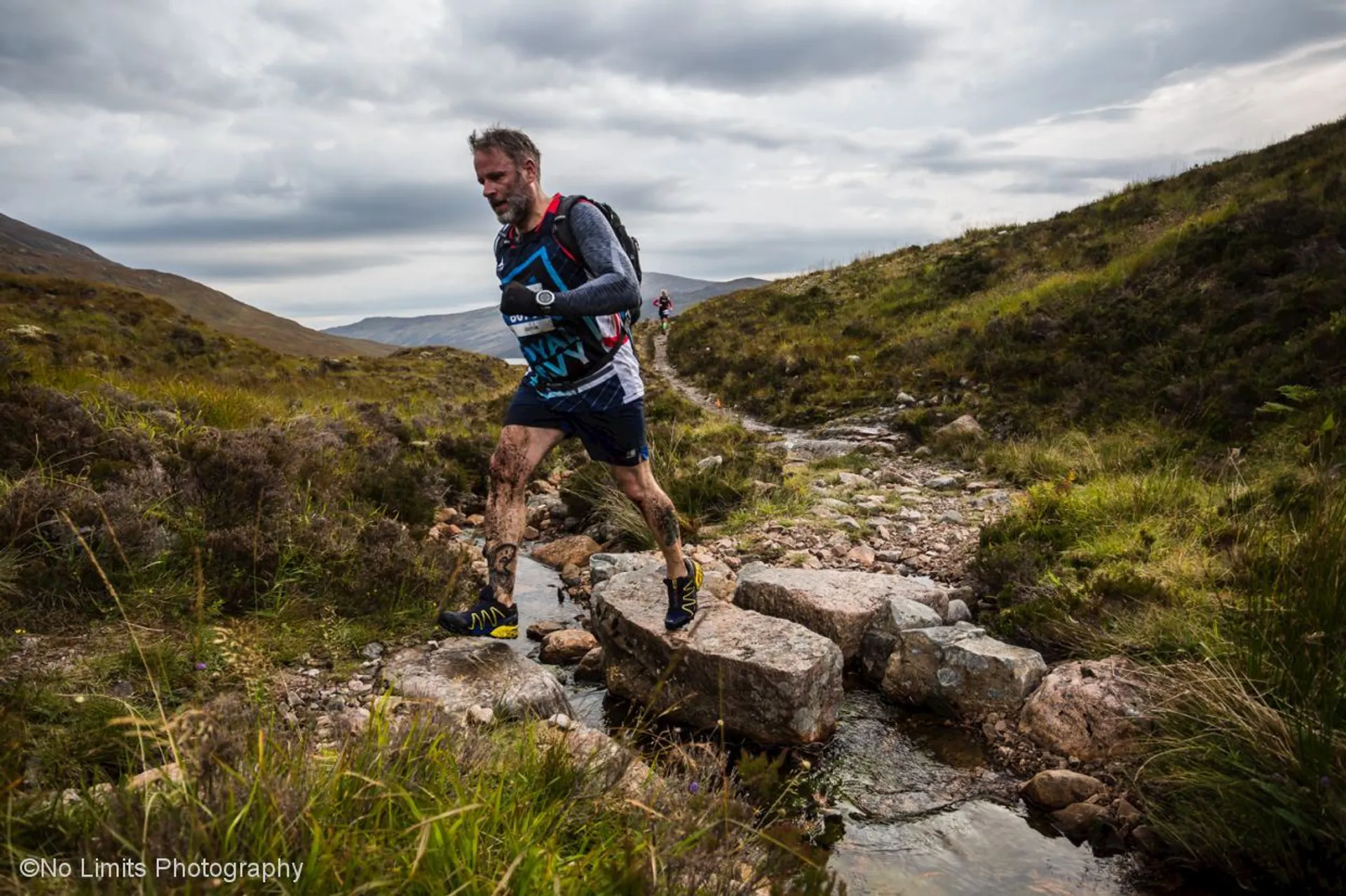 The image shows a man trail running amidst a natural landscape. He appears to be