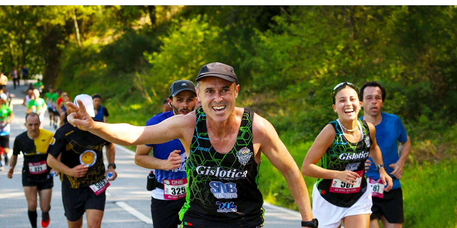 This image depicts a group of runners participating in a road race or marathon. The person in the foreground, presumably a man, is smiling broadly and reaching out toward the camera, giving off an enthusiastic and energetic vibe. He is wearing a hat, sunglasses, and a tank top that suggests sponsorship or team affiliation, and has a race number attached to his shorts, indicating his registration in the event. Other runners in the background also appear focused and determined, each at different stages in their stride. The environment suggests a sunny day with a route that takes them through a natural, verdant setting, likely providing a picturesque backdrop for the race.