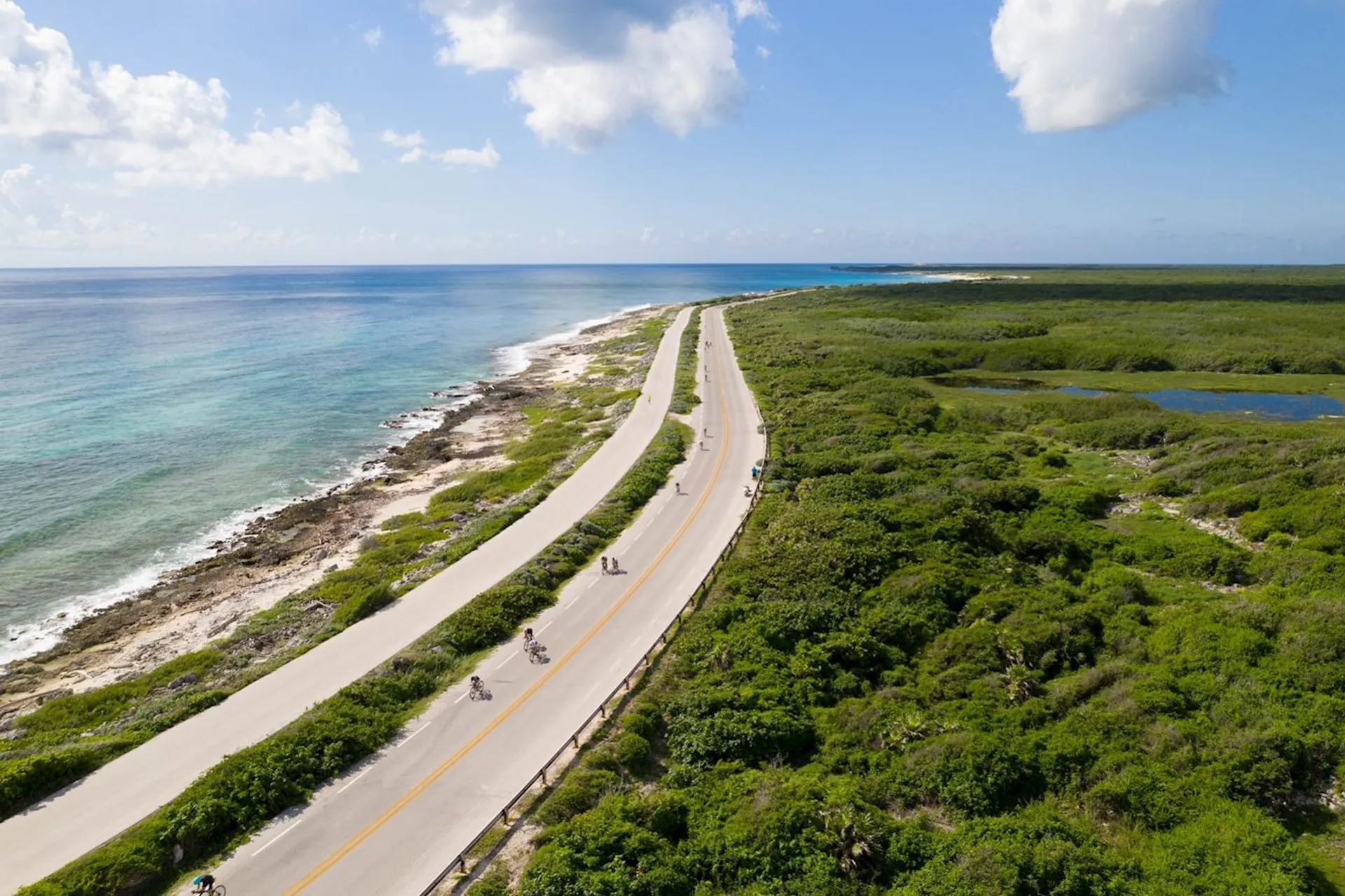 The image shows a beautiful aerial view of a long, straight road that stretches between two contrasting landscapes. On one side of the road, there's a tranquil beach and the clear blue sea, while on the other side, there appears to be a dense green vegetation area which could be a marsh or mangrove. Something that stands out is the lack of development aside from the road, which suggests this area may be preserved or less inhabited. On the road, there are cyclists riding in single file, enjoying what must be a scenic route. It looks like an ideal place for a peaceful drive or cycle with stunning natural views on either side.