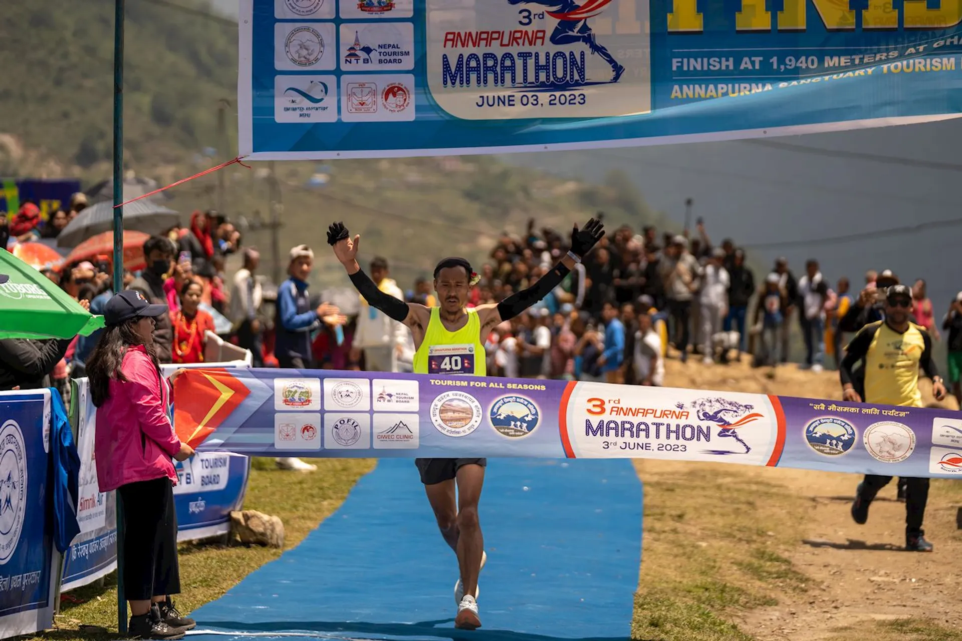 The image depicts a runner crossing the finish line of the 3rd Annapurna Marathon. The finish line banner displays the marathon's name and date, June 3, 2023. There are spectators and various event signs around the finishing area.