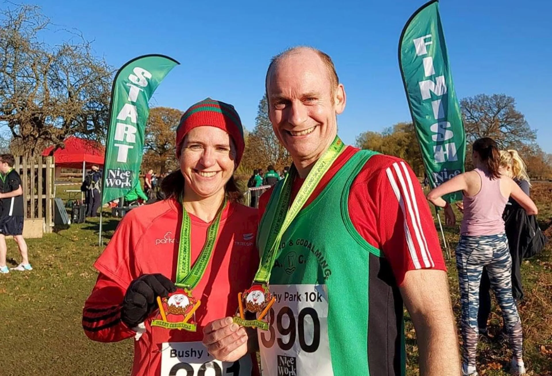 The image shows two individuals standing outdoors, likely at a running event. They are both wearing athletic attire and bibs with numbers, indicating they have participated in a race. The man on the right is wearing a green tank top and the woman on the left is wearing a red long-sleeve shirt; both appear to be in good spirits. They are each holding a medal, suggesting they have completed the race and possibly received awards for their performance. Behind them are banners with the words "Start" and "Finish," consistent with start/finish lines of a race, and there are other participants and spectators in the background. The setting looks like a park, and it appears to be a sunny day, which contributes to the cheerful atmosphere of the event