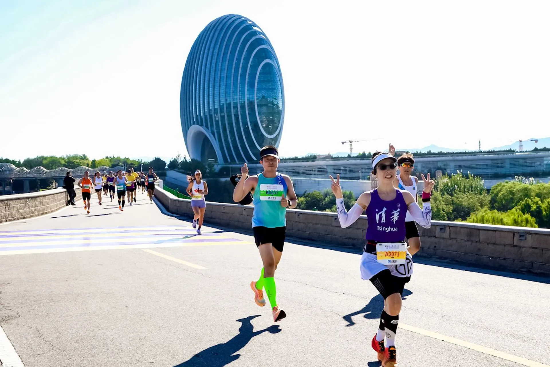 The image shows a group of people running a race on a road. In the background, there is a modern, oval-shaped building with a distinctive architectural design. The runners appear to be participating in an organized event, wearing numbered bibs and athletic gear. The setting is outdoors, with a clear sky and some vegetation visible.