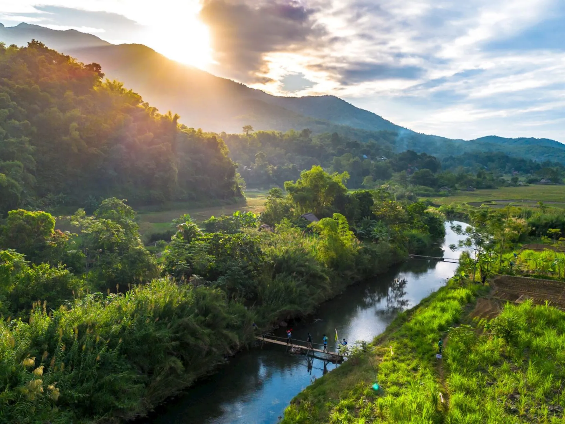 The image depicts a scenic landscape with a river running through a lush, green area. There are hills or mountains in the background partially illuminated by the setting or rising sun, casting a warm glow across the scene. A small footbridge crosses the river, and there are a few people on it, surrounded by dense vegetation. The sky is partly cloudy, adding to the serene and picturesque atmosphere.