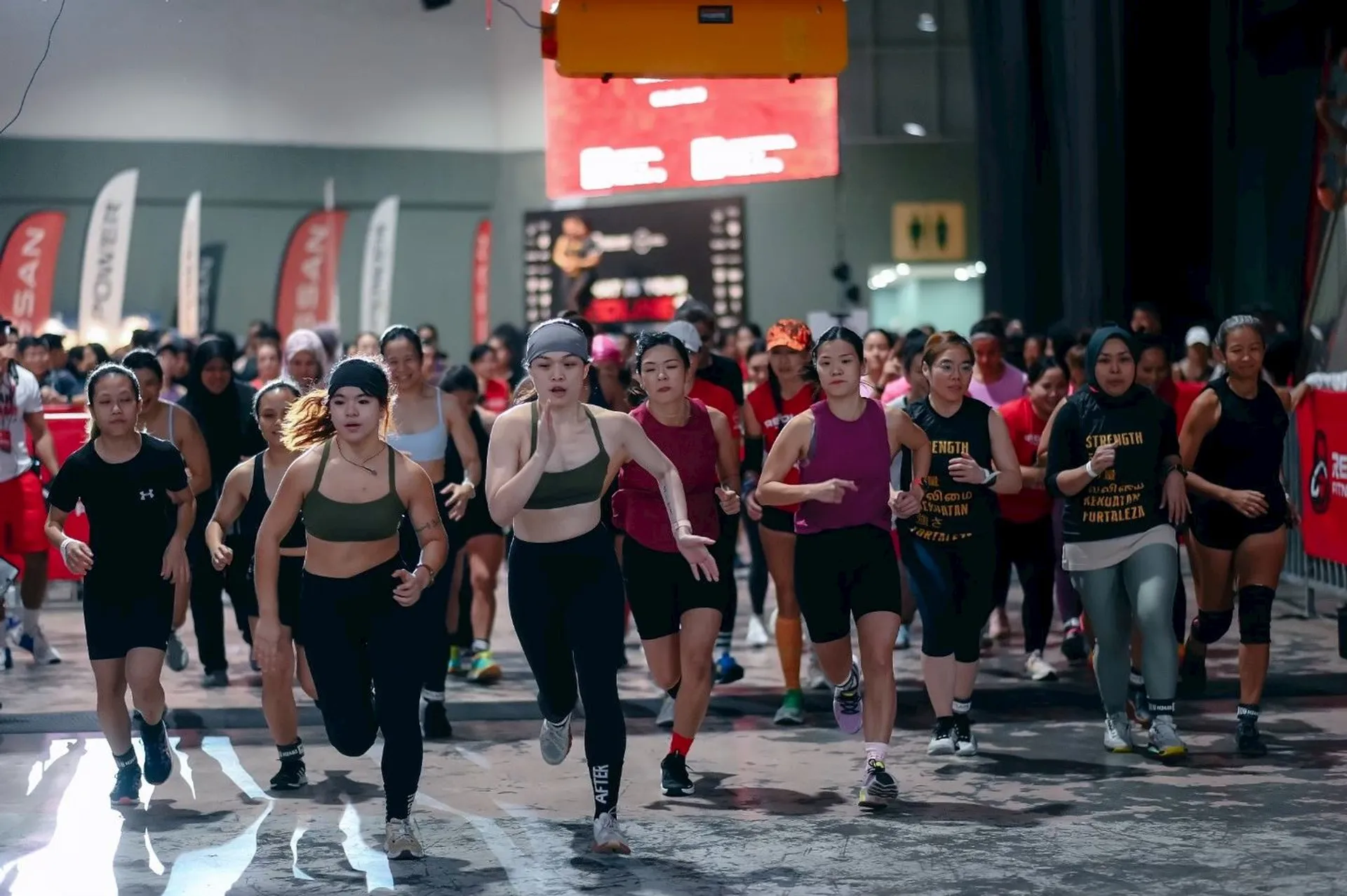 The image shows a group of people participating in a race or athletic event indoors. They appear to be running together, and there are banners and signs in the background, indicating it might be an organized sporting event.