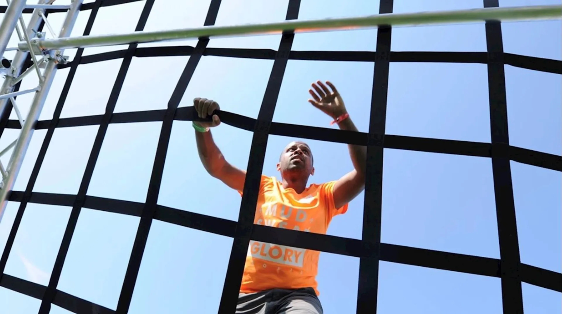 The image shows a person climbing a net or rope structure against a clear blue sky. The person is wearing an orange shirt and appears to be participating in an outdoor challenge or obstacle course.