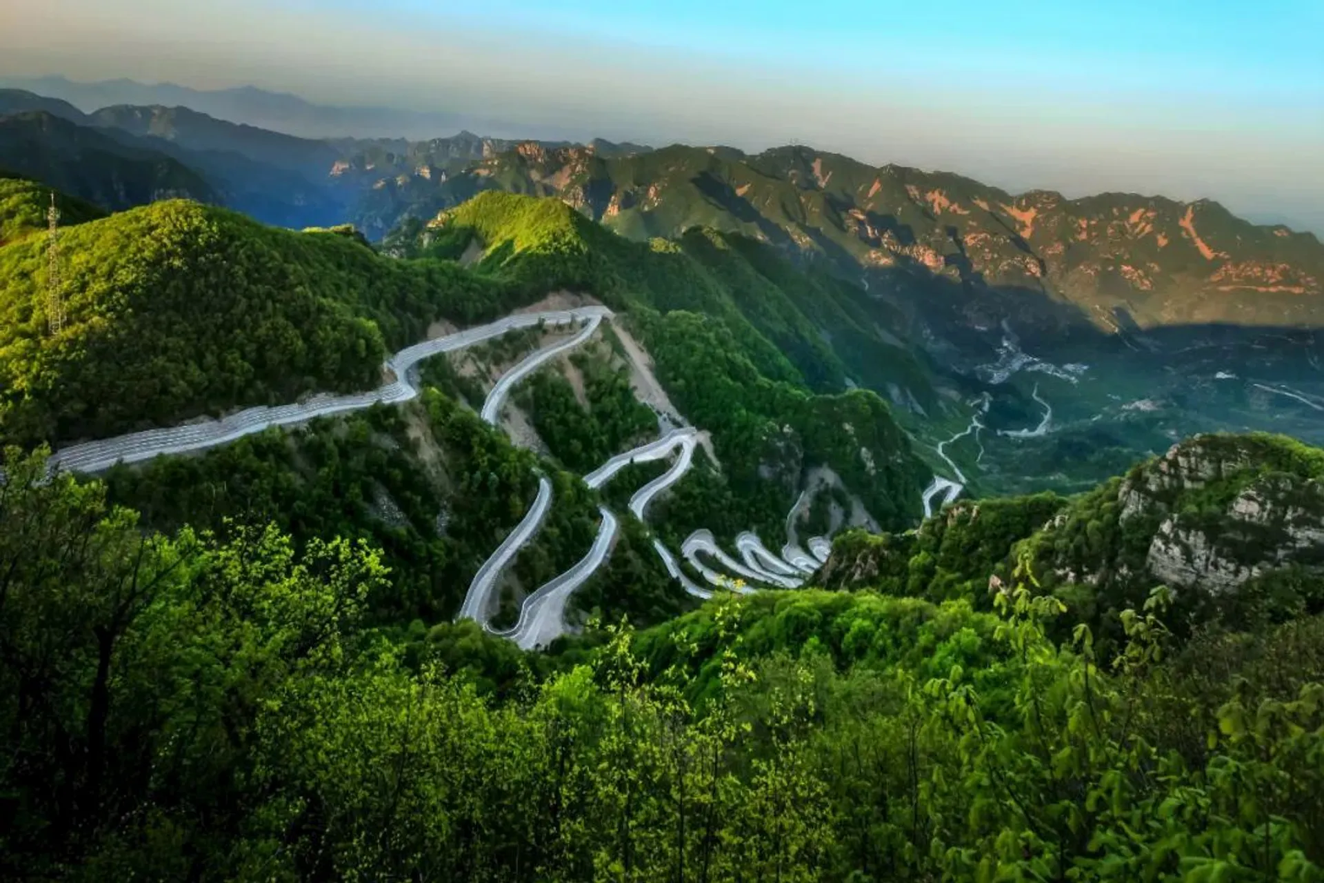 This image depicts a scenic landscape of a winding mountain road surrounded by lush green hills and valleys. The road curves and loops through the mountains, offering a striking view of the natural landscape. The background features more mountains under a clear blue sky, creating a serene and picturesque scene.