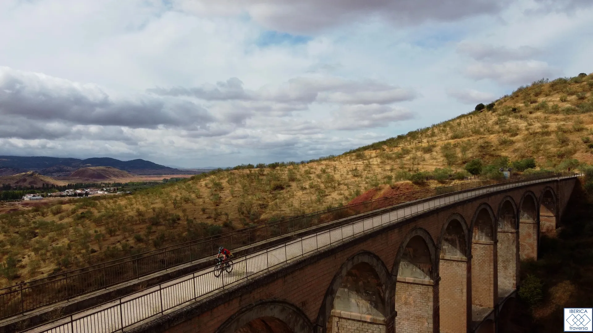 The image features a cyclist riding on a long, elevated bridge or viaduct with several arches. The landscape includes hills covered with shrubs and grass, and there's a cloudy sky overhead.