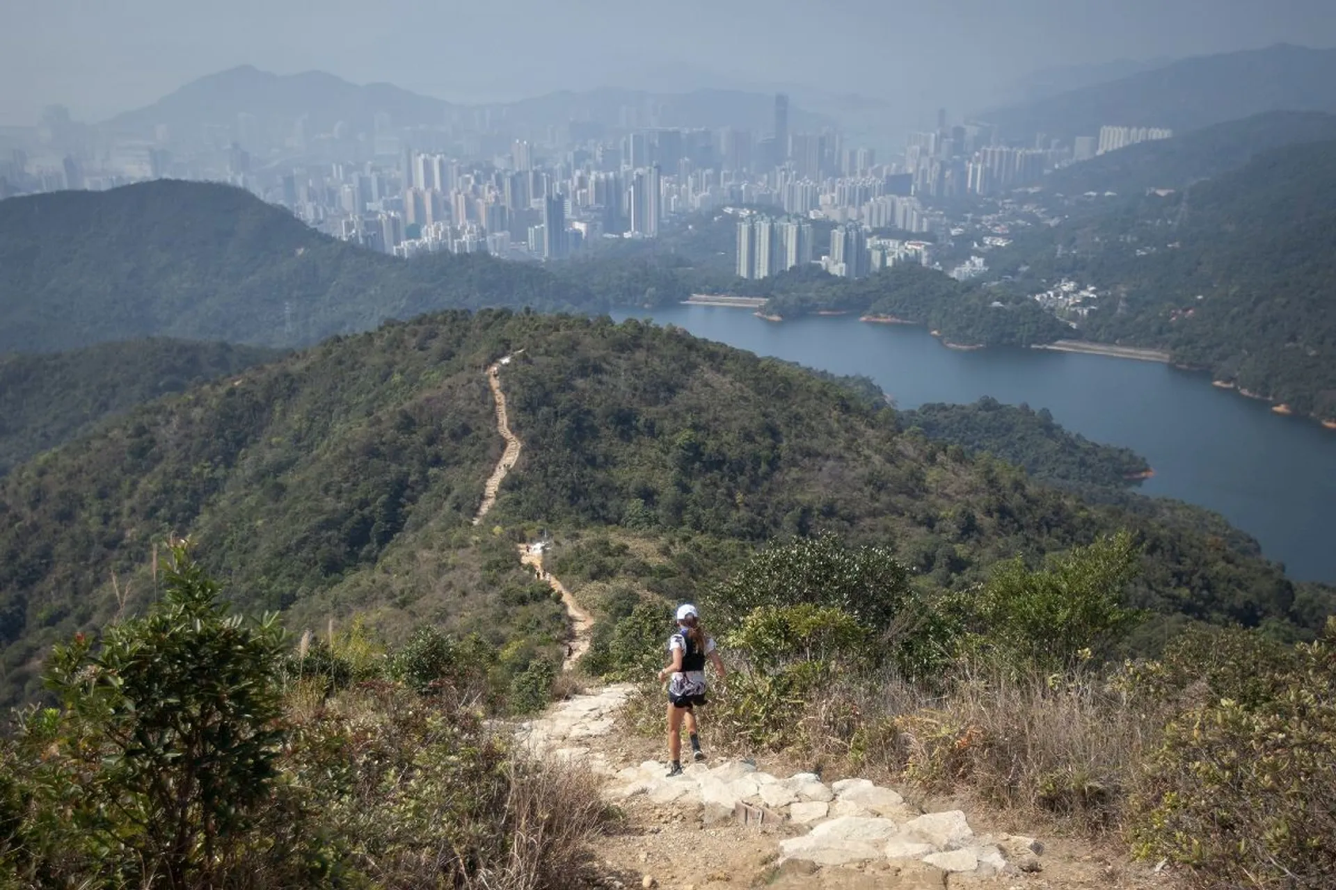 The image shows a landscape view from a hiking trail. The foreground features a hiker walking along a path surrounded by greenery. In the background, there is a cityscape with numerous buildings, and a body of water is visible between the hills. The scene is set in a mountainous area, suggesting a blend of natural and urban environments.