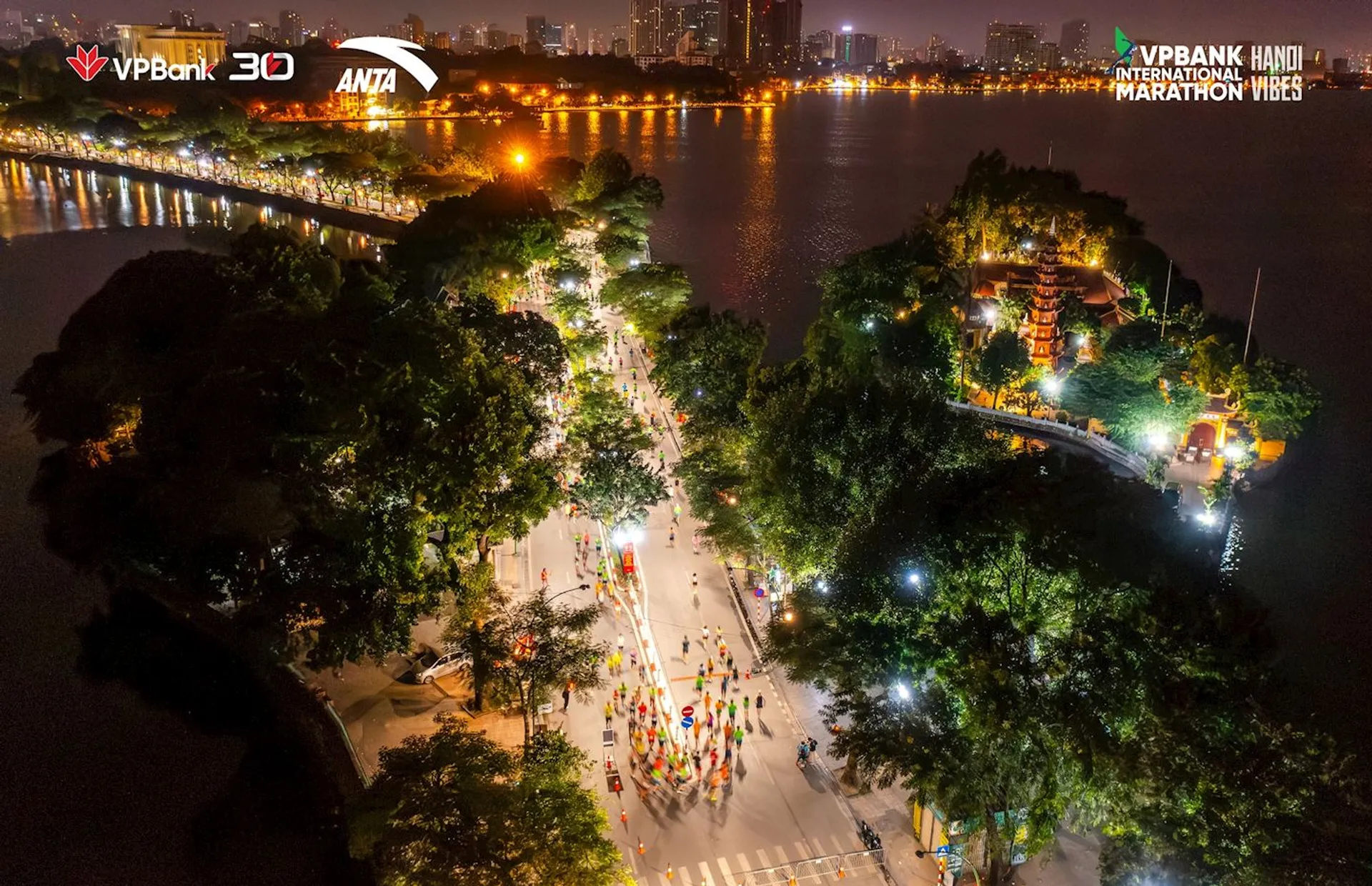 This image depicts a vibrant nighttime scene with a busy road or pathway alongside a body of water. There seem to be many people gathered along the path, possibly participating in an event or activity such as a marathon, as indicated by the presence of banners with logos and what may be the word "MARATHON". There are colorful features which might be decorative lights or installations among the people. The area is well-lit and appears festive or celebratory. Surrounding the area are trees and what looks like a park setting, with buildings illuminated in the background, which suggests an urban environment. The vantage point suggests the photo was taken from an elevated position or a drone. The lettering in the top corners of the image represents branding or sponsorships,