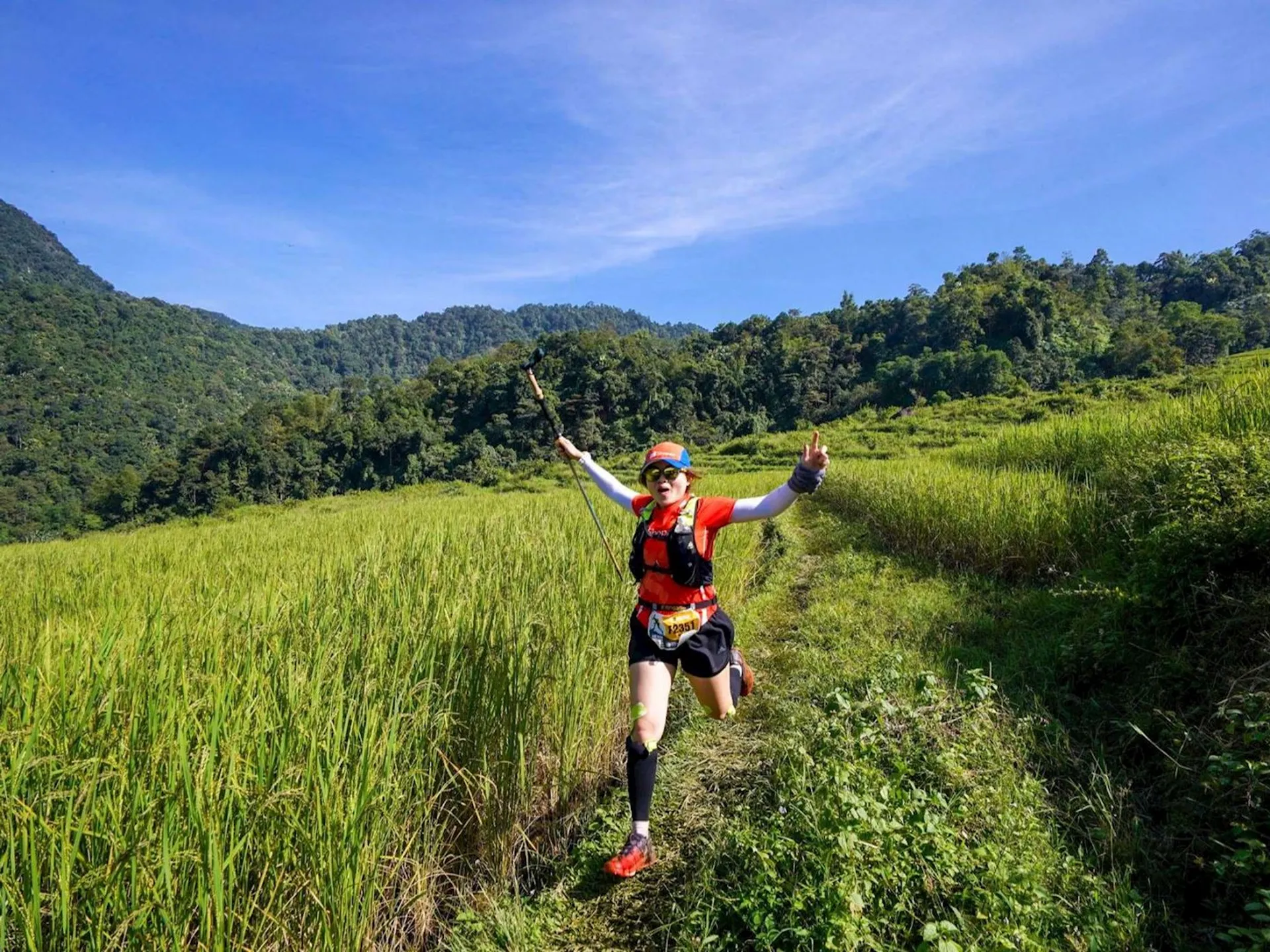 The image shows a person running energetically along a path in a lush, green landscape. The person is dressed in sporty attire, including a red shirt, shorts, and a cap, and is carrying what appears to be a selfie stick. They are surrounded by tall grass or crops, with a backdrop of hills and a clear blue sky. The scene suggests a joyful and active outdoor experience, possibly during a trail run or similar activity.