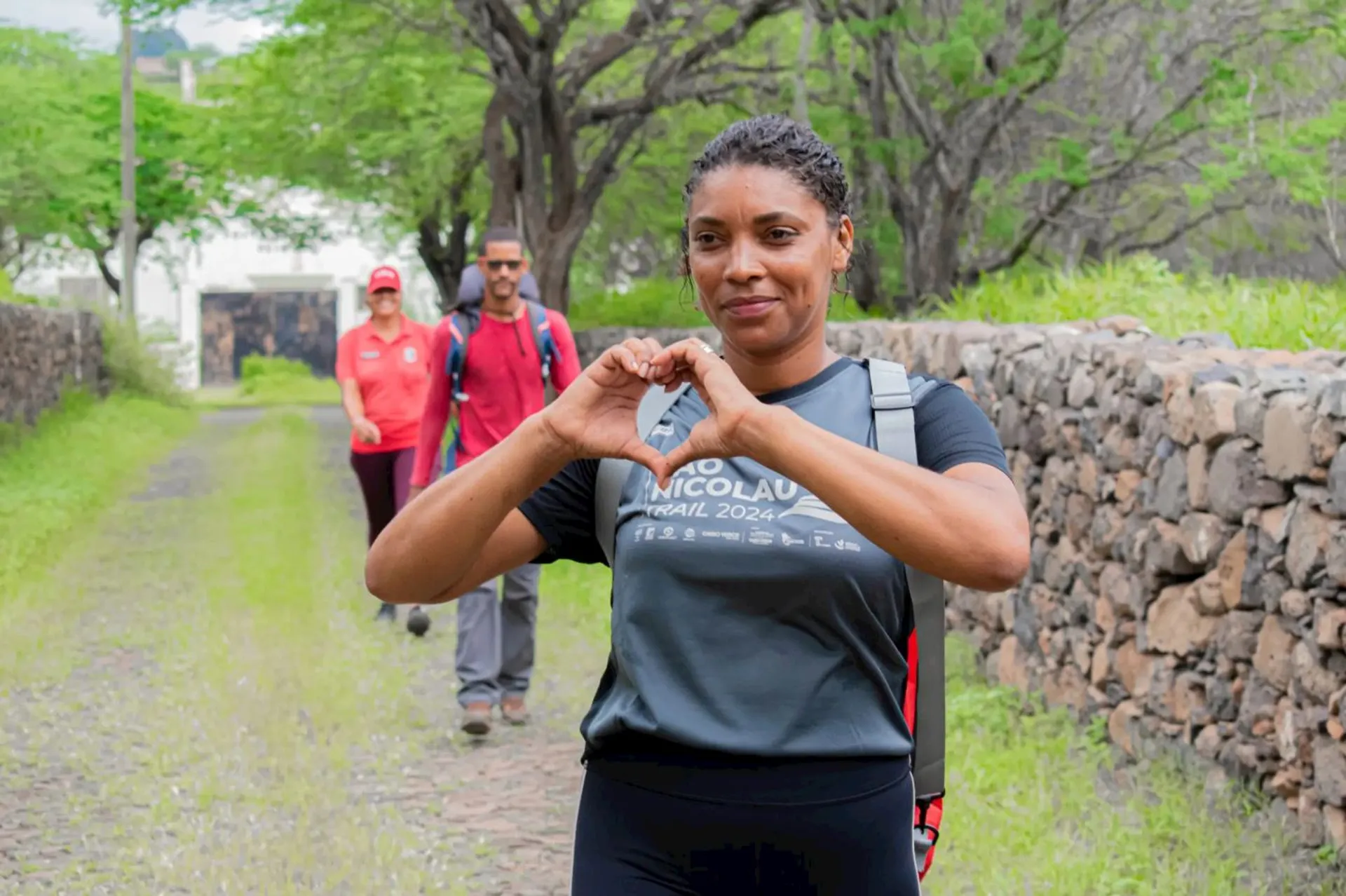The image shows a woman making a heart shape with her hands, walking on a path lined with stone walls and greenery. Behind her, two other people are walking. The scene appears to be outdoors, possibly in a rural or natural setting.