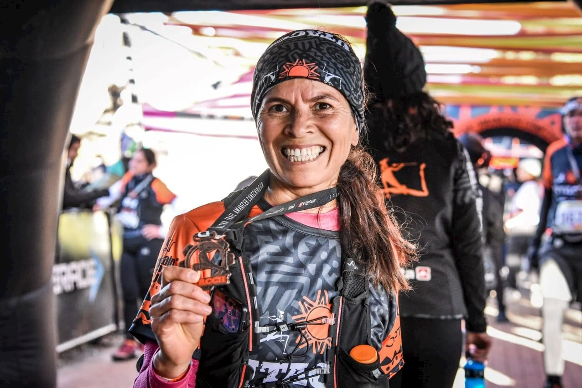 The image shows a person smiling and holding up a medal, possibly having just completed a race or sporting event. They are wearing athletic gear and a headband. Other people and event-related structures are visible in the background.