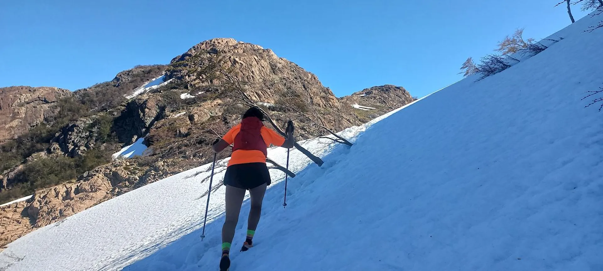 The image shows a person hiking up a snowy mountain slope. They are wearing an orange top and black shorts, and using poles for support. The background includes rocky terrain and a clear blue sky.