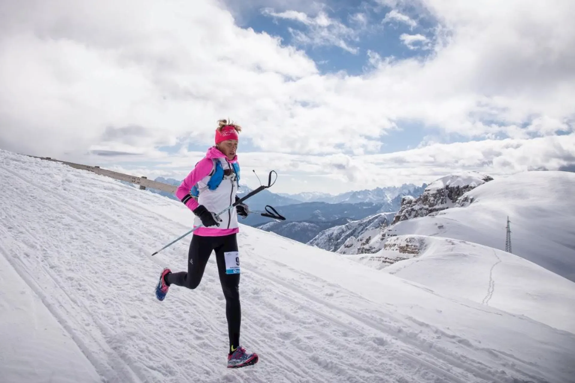In the image, there is a person engaged in a winter sporting activity, specifically Nordic skiing or ski mountaineering. The individual is dressed in athletic attire suitable for cold weather, including a pink long-sleeve top, black leggings, gloves, and a pink headband. They are using specialized lightweight skis and poles designed for ascending and traversing snowy terrain. The background showcases a breathtaking snowy mountain landscape with a mix of overcast and blue skies, suggesting the setting is at a high altitude where such recreational activities are common.