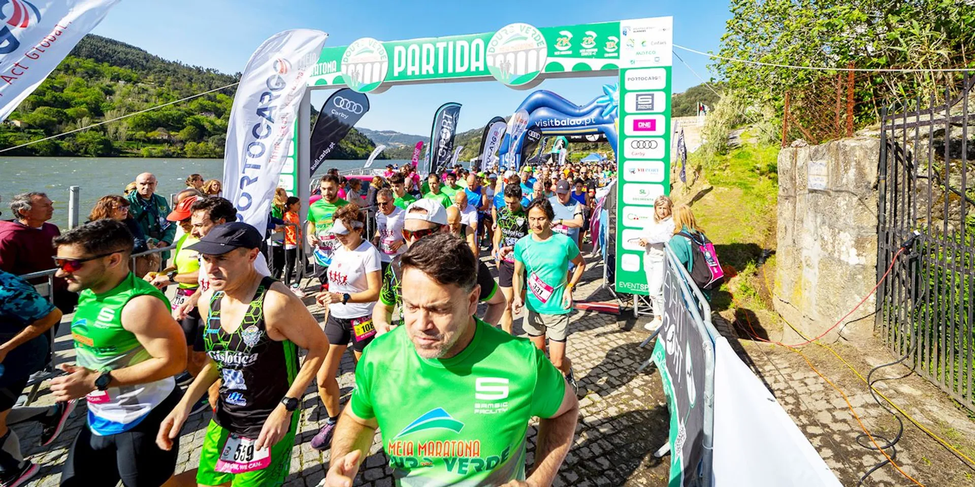 The image shows a group of runners at the start line of a running event, ready to embark on their race. The setting appears to be in a natural area with greenery and a body of water in the background, suggesting it might be a scenic route. Several banners and inflatable arches indicate sponsorship and mark the starting point, labeled "PARTIDA" which means "start" in Portuguese or Spanish. The participants are wearing athletic clothing and race bibs, signifying it's an organized sporting event. The atmosphere looks vibrant and energetic, with everyone poised to begin their run.