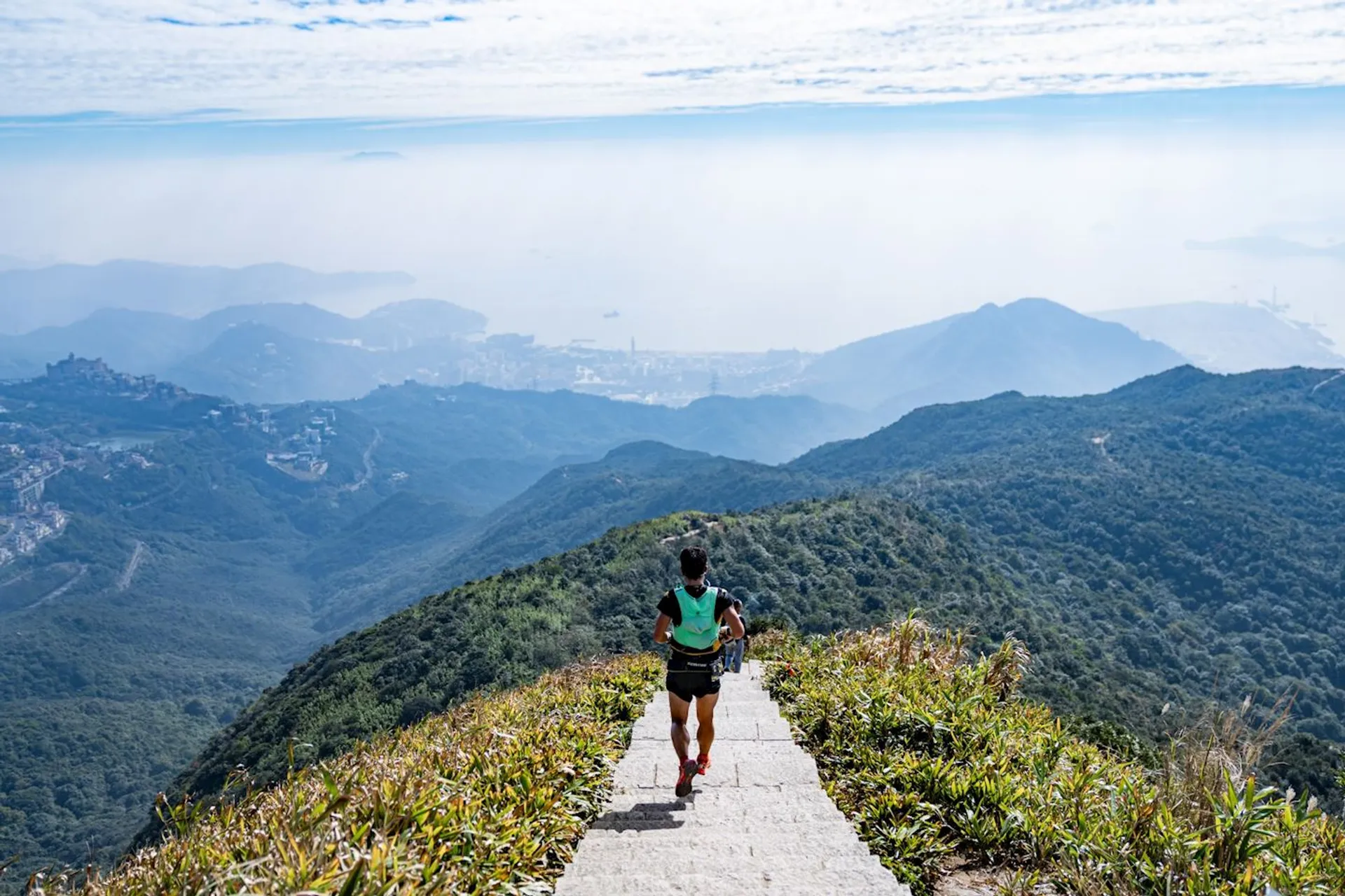 The image shows a person running on a paved path leading down a hill or mountain. The runner is wearing a dark-colored top, shorts, and red running shoes. The surrounding landscape features lush green hills or mountains under a clear sky with scattered clouds. In the distance, there appears to be a cityscape or urban area with buildings visible through a slight haze, which suggests the photo might have been taken from a high vantage point, overlooking the city or town below. The overall scene conveys a sense of tranquility, exercise in nature, and the beauty of outdoor landscapes.