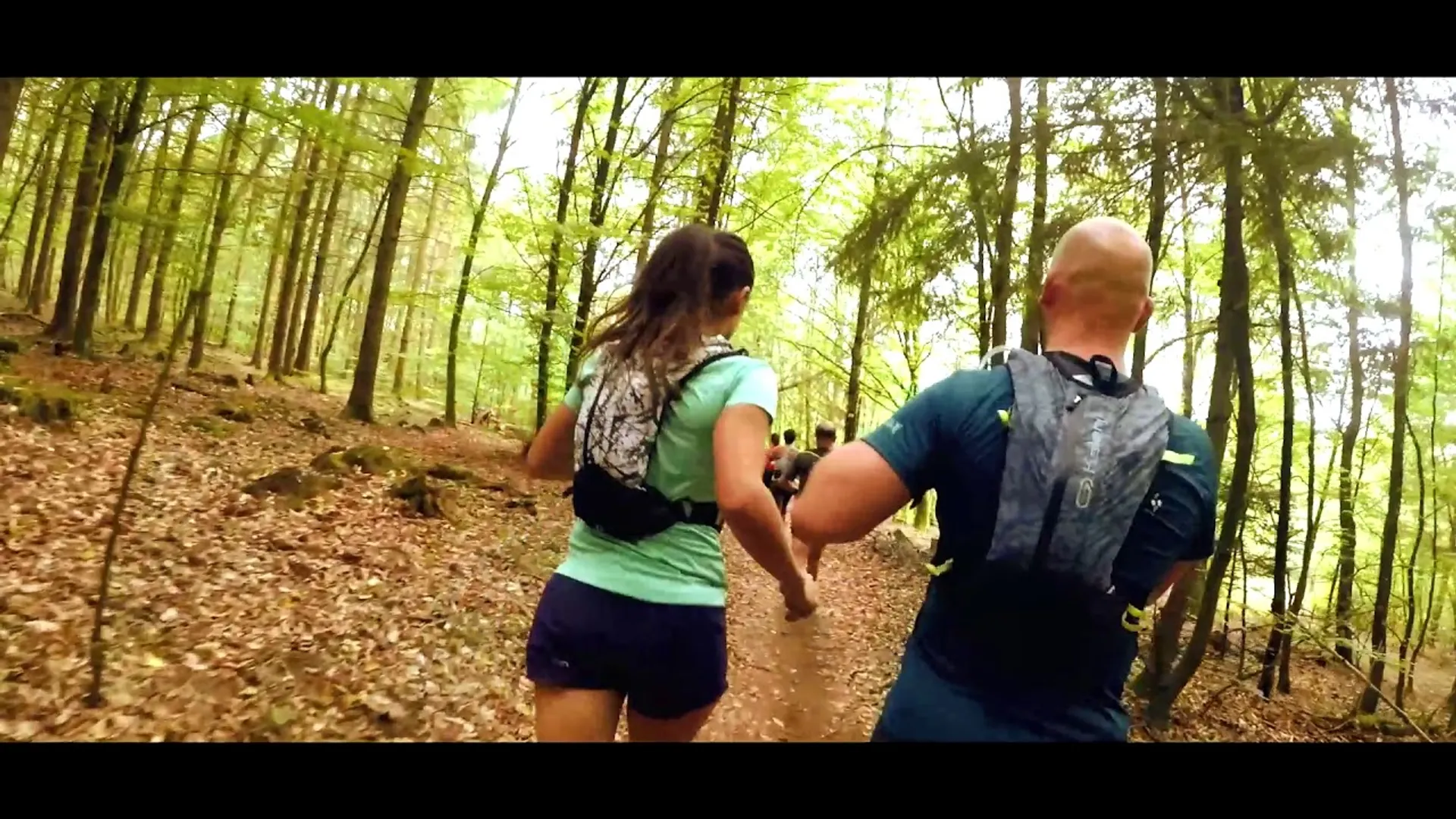 The image shows two people running together on a forest trail. Both runners are wearing
