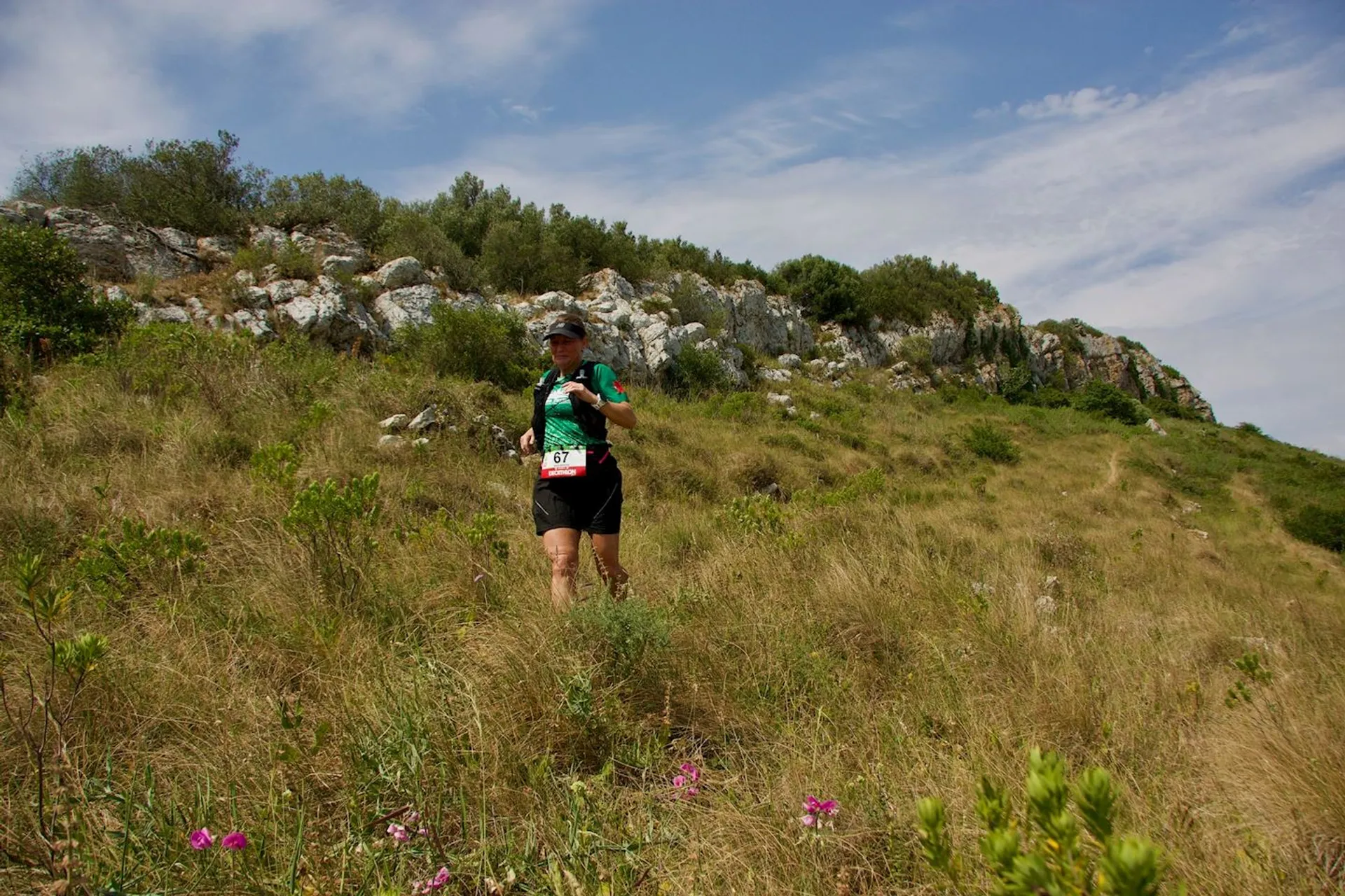 The image shows a person running or hiking on a grassy and rocky hillside. The individual is wearing athletic gear, including a shirt and shorts, and has a race bib. The sky is partly cloudy, and there is a rocky formation in the background. Purple wildflowers are visible in the foreground.