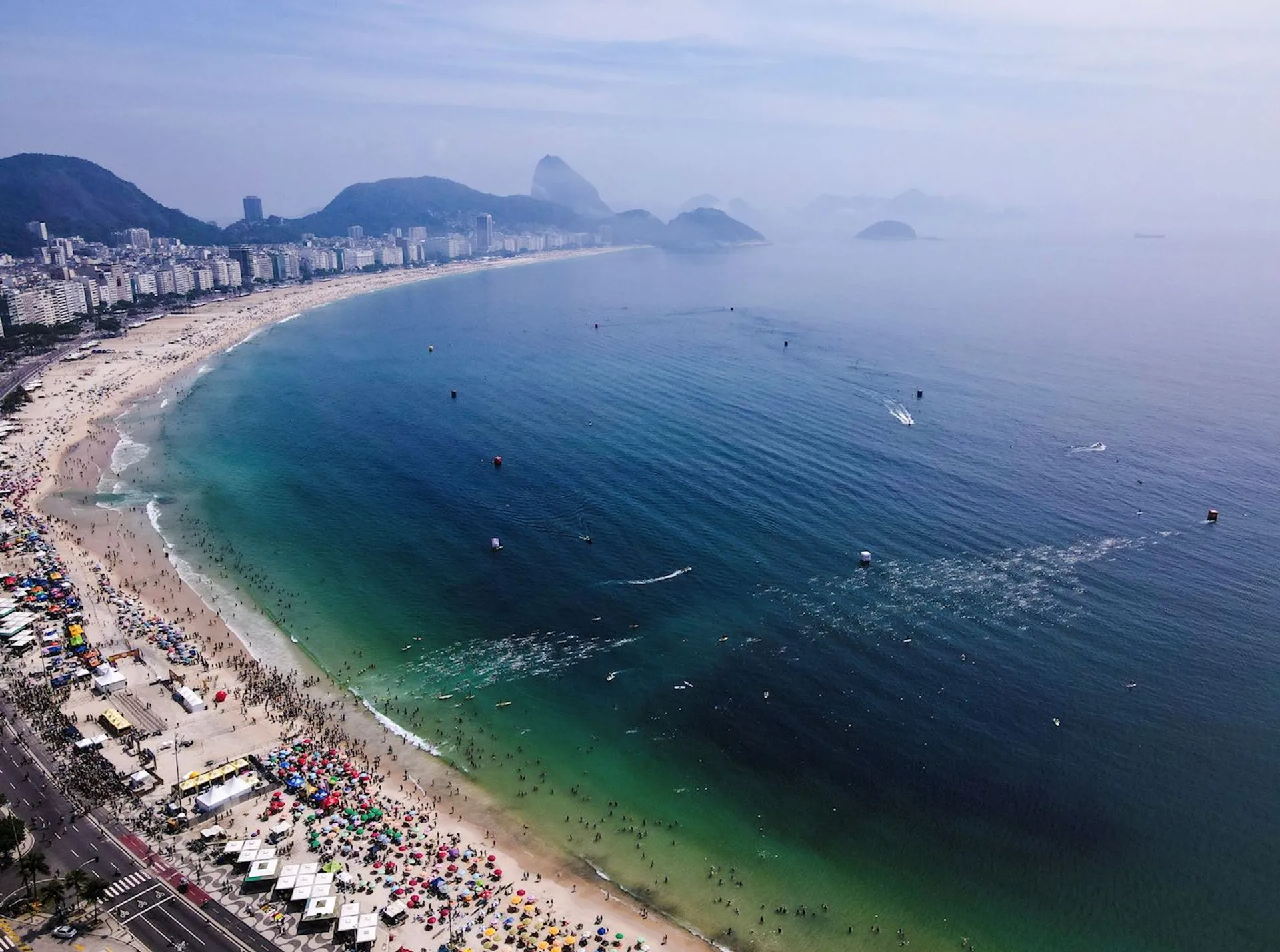 The image shows a scenic view of a beach with a curved shoreline, likely Copacabana Beach in Rio de Janeiro, Brazil. The beach is bustling with people, umbrellas, and beachgoers along the shoreline. In the background, you can see buildings, and beyond them, mountains and a hazy sky. The ocean water is dotted with some boats and swimmers.