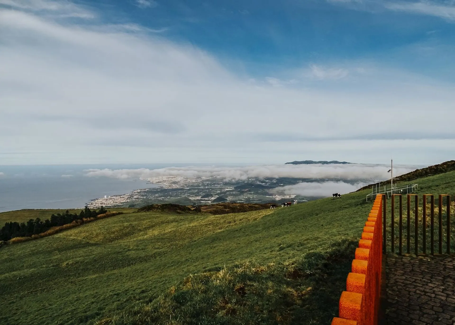 The image depicts a scenic landscape view from a high vantage point. In the foreground, there is a grassy slope with a distinct orange-colored path or edge, alongside a railing or fence possibly for safety. The middle ground shows a lush area that gently slopes down towards a coastal region, with low-lying clouds or fog hovering above the sea, giving a serene and somewhat ethereal appearance to the scenery. In the background, you can see the outline of a mountain or island, adding depth to the scene. The sky is clear with only a few scattered clouds, suggesting a peaceful day with good visibility. There are no people or animals visible in this image.