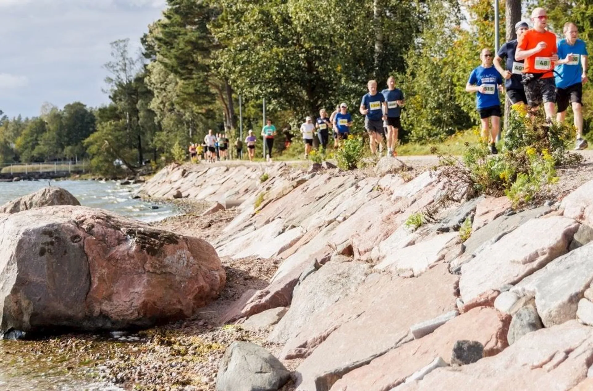 The image shows a group of people running on a path next to a body of water. The runners are wearing athletic clothing, and there are trees and rocks alongside the path. It appears to be a scenic area, possibly a park or waterfront trail.