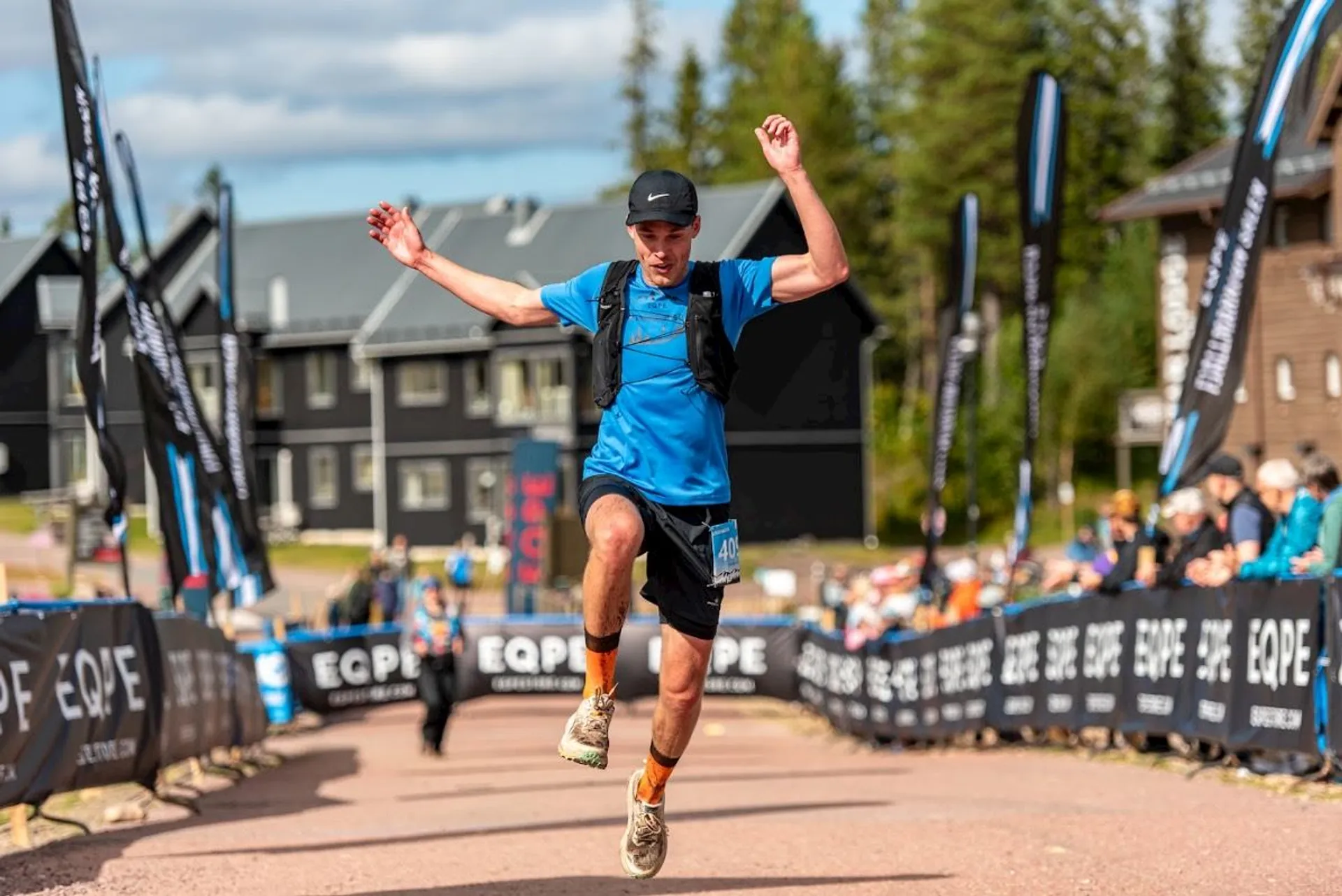 The image shows a person running a race. They are in mid-air, appearing to be celebrating or finishing. The person is wearing athletic gear, including a cap, a vest, and a blue shirt. There are banners along the race path and spectators in the background. The setting appears to be outdoors, with buildings and trees visible.