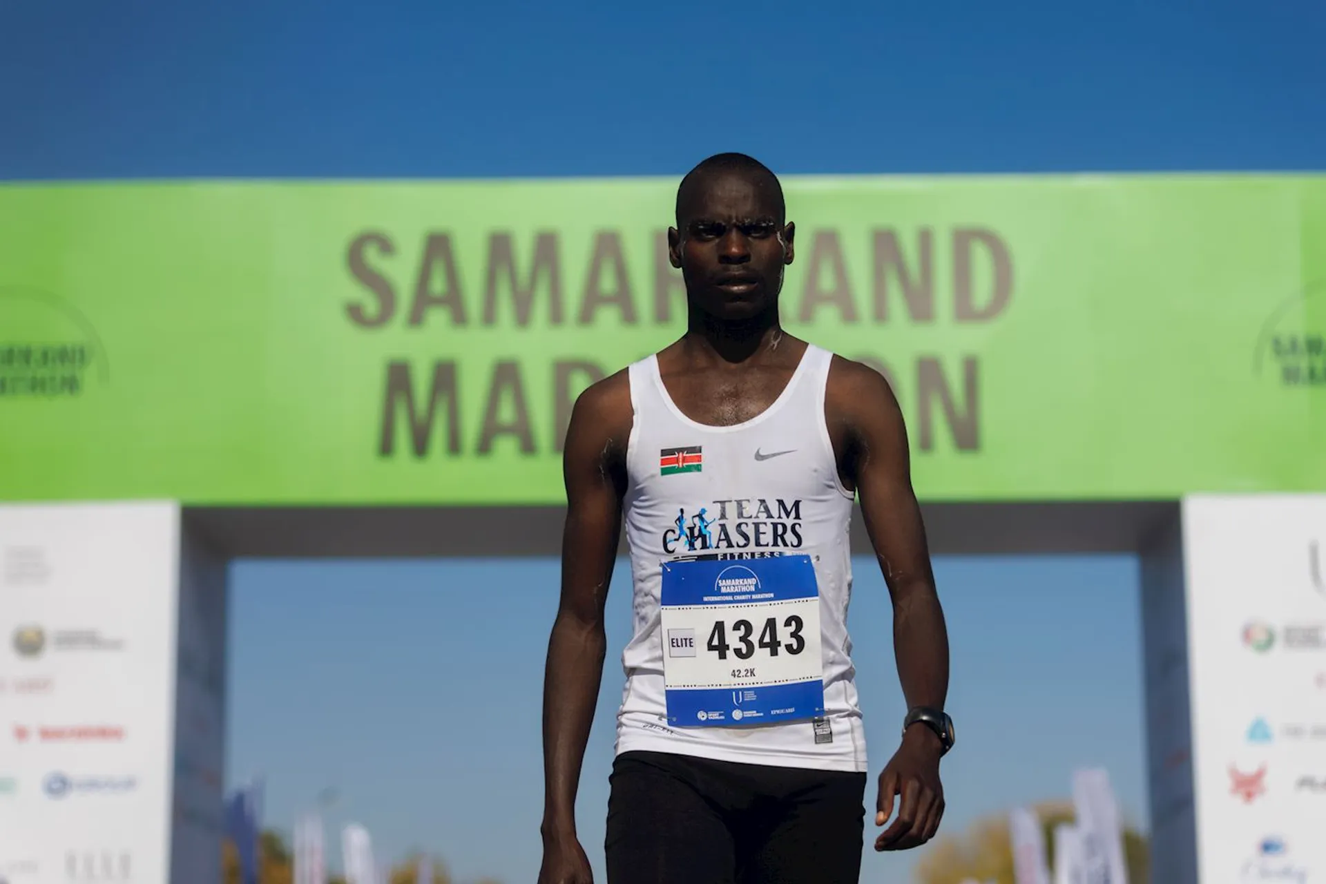 The image shows a runner wearing a white tank top with "Team Chasers" on it, along with a number bib (4343) over a blue backdrop. He's standing in front of a finish line or starting line with a green banner that says "Samarkand Marathon."
