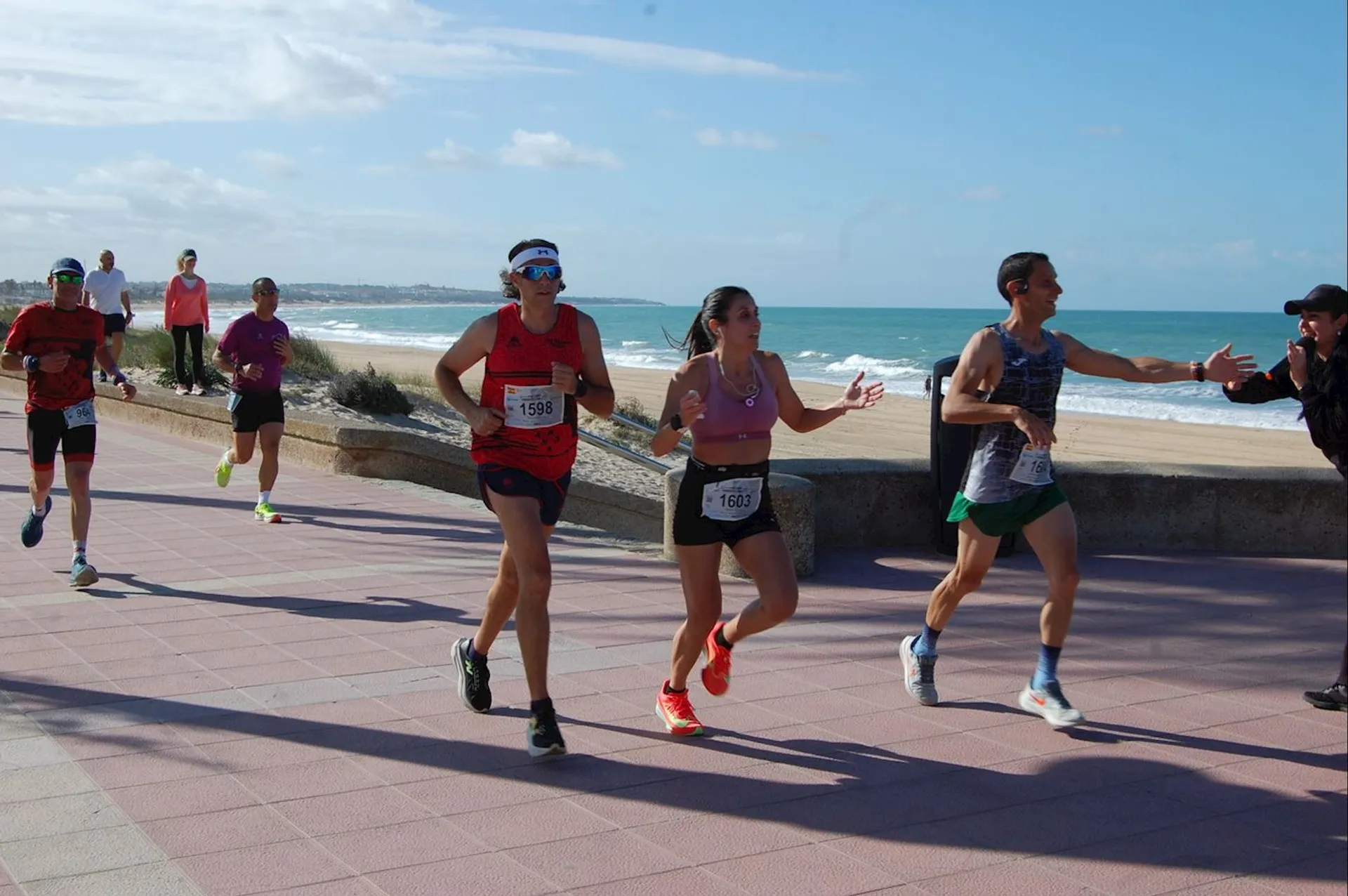 The image shows a group of people running along a seaside pathway. They appear to be participating in a race, as they are wearing running gear and bib numbers. The background features a beach and the ocean. A few people are observing or interacting with the runners.