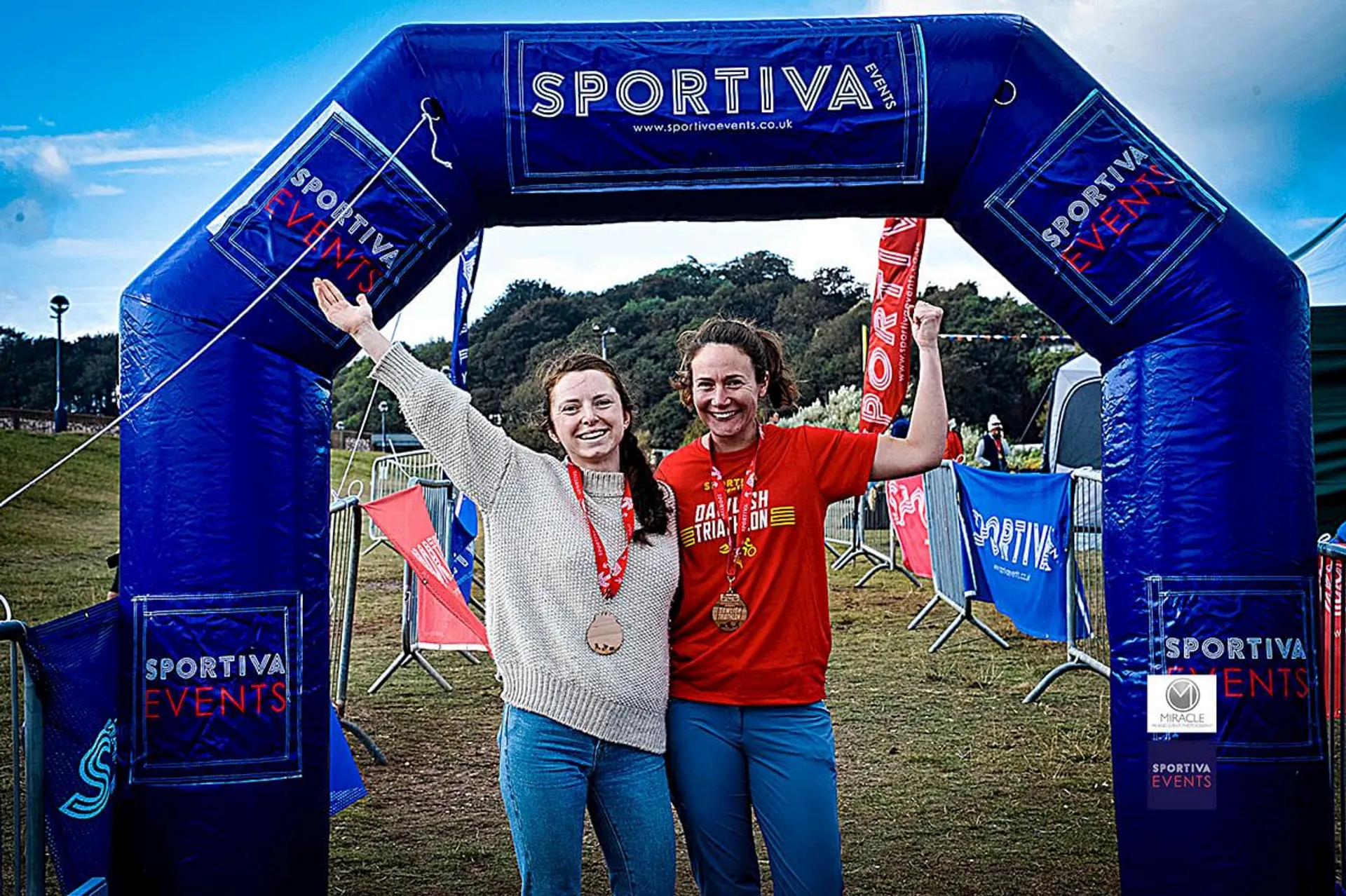 The image shows two people smiling and standing under an inflatable arch with the text "